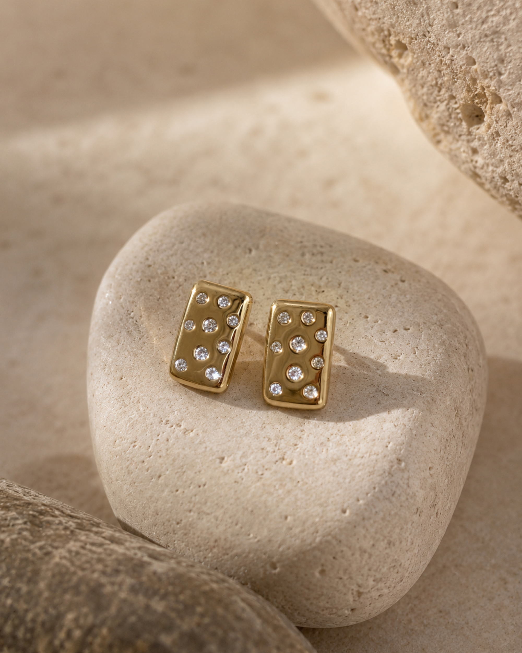 A pair of gold rectangular stud earrings with small clear stones, displayed on a smooth beige stone with rough natural stones in the background.