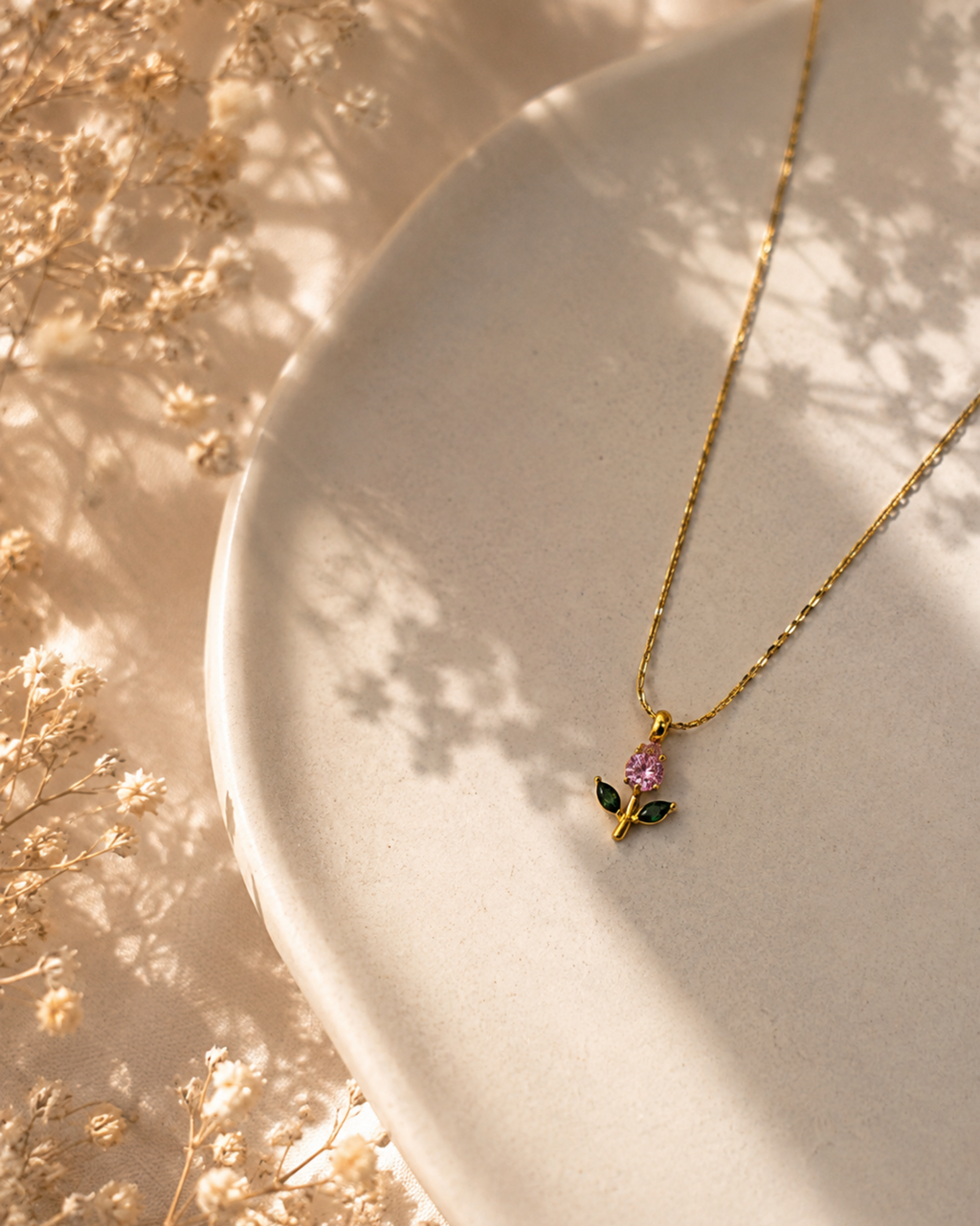 A delicate gold chain necklace with a small flower pendant featuring a pink center and green leaves, displayed on a textured beige ceramic plate with soft dried floral decorations and dappled sunlight.
