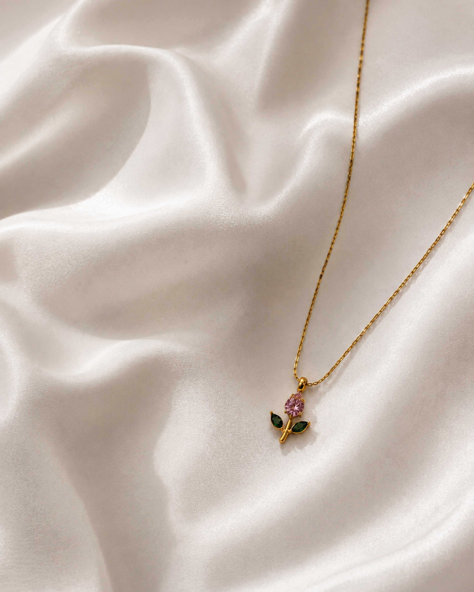A delicate gold chain necklace with a small pendant shaped like a flower, featuring a pink gemstone and green enamel leaves, displayed on draped white satin fabric.