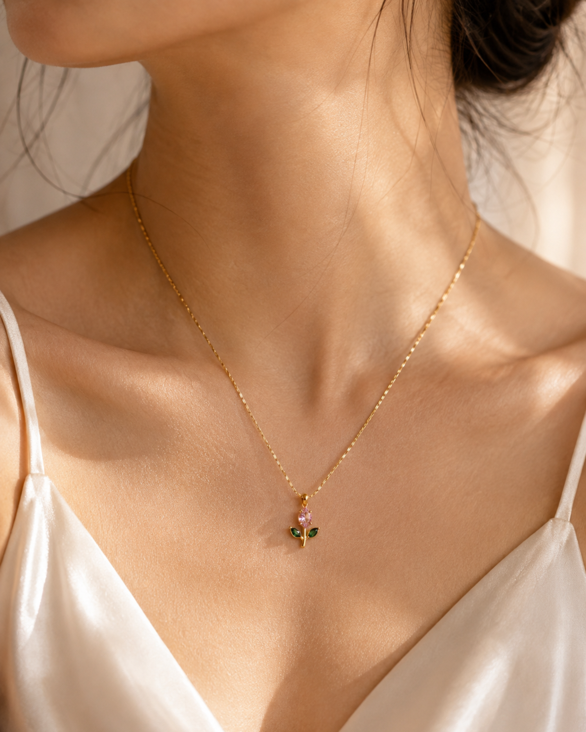 A close-up of a woman's neck and upper chest wearing a delicate gold chain necklace with a small clover-shaped pendant featuring green, pink, and gold details, paired with a white satin camisole.