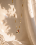 A delicate gold chain necklace with a small pendant featuring a pink teardrop-shaped stone and green leaves, displayed against a softly draped cream fabric background with warm sunlight and shadows.