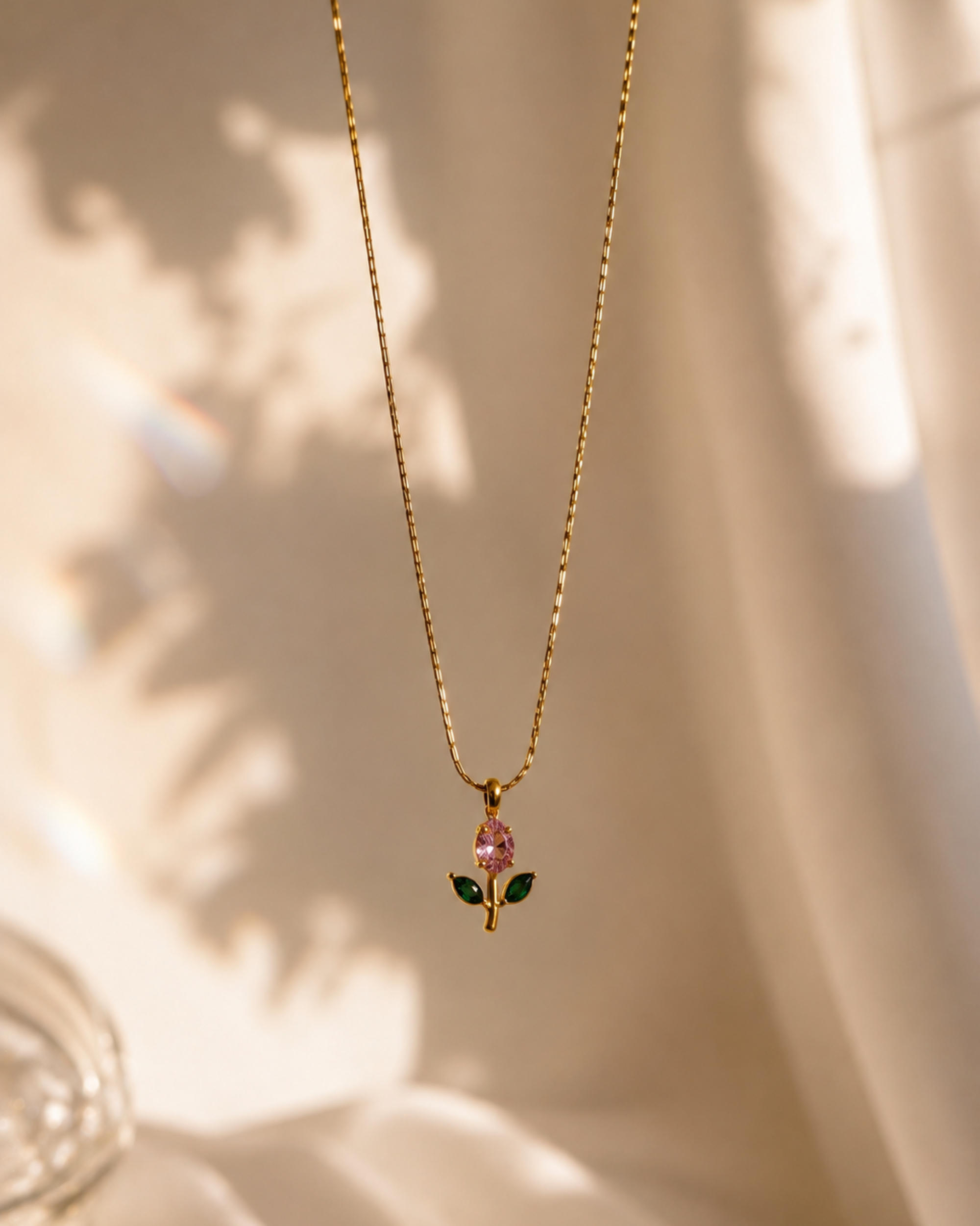 A delicate gold chain necklace with a small pendant featuring a pink teardrop-shaped stone and green leaves, displayed against a softly draped cream fabric background with warm sunlight and shadows.