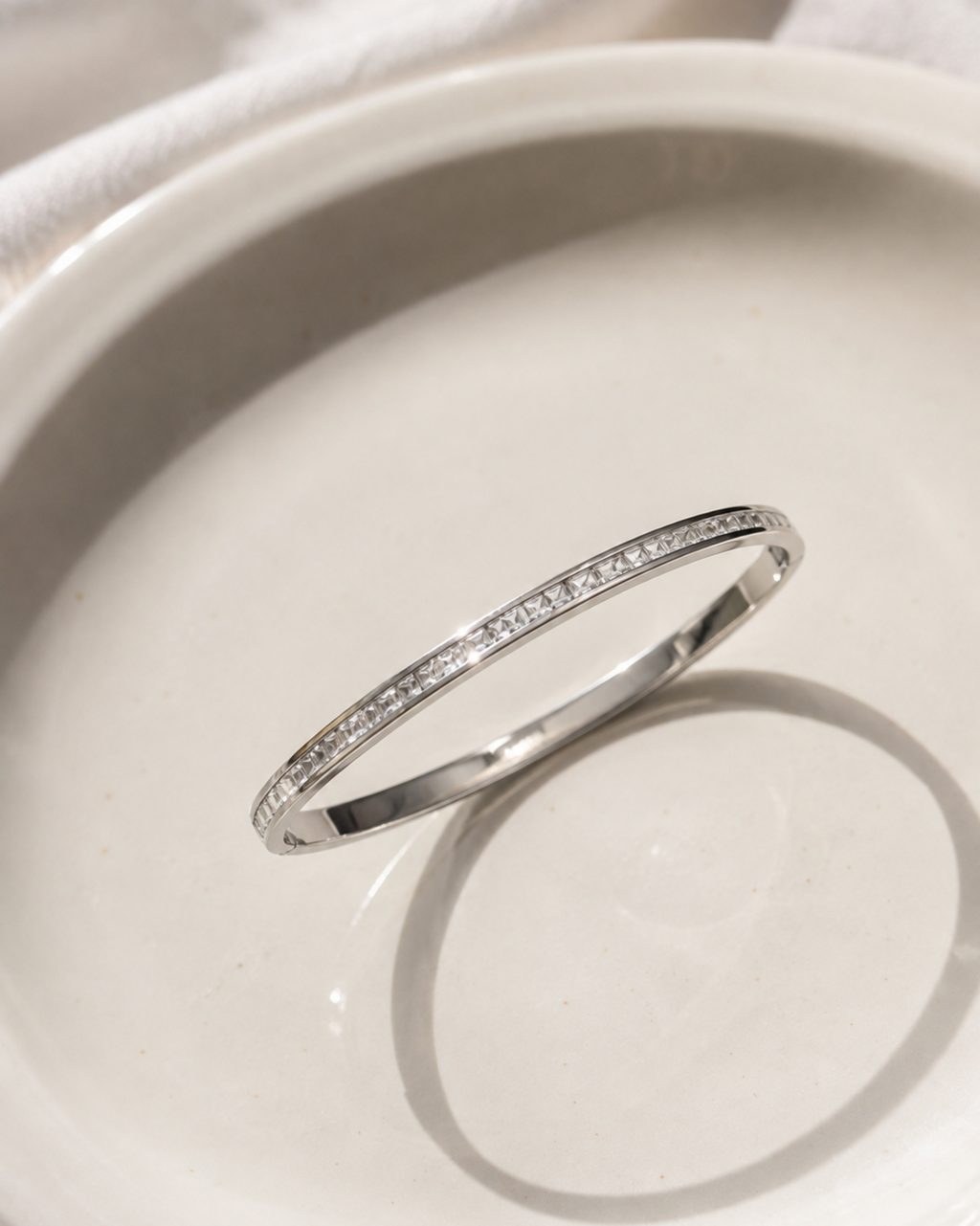 A thin silver band ring with a row of small clear stones, photographed resting on a white ceramic dish.