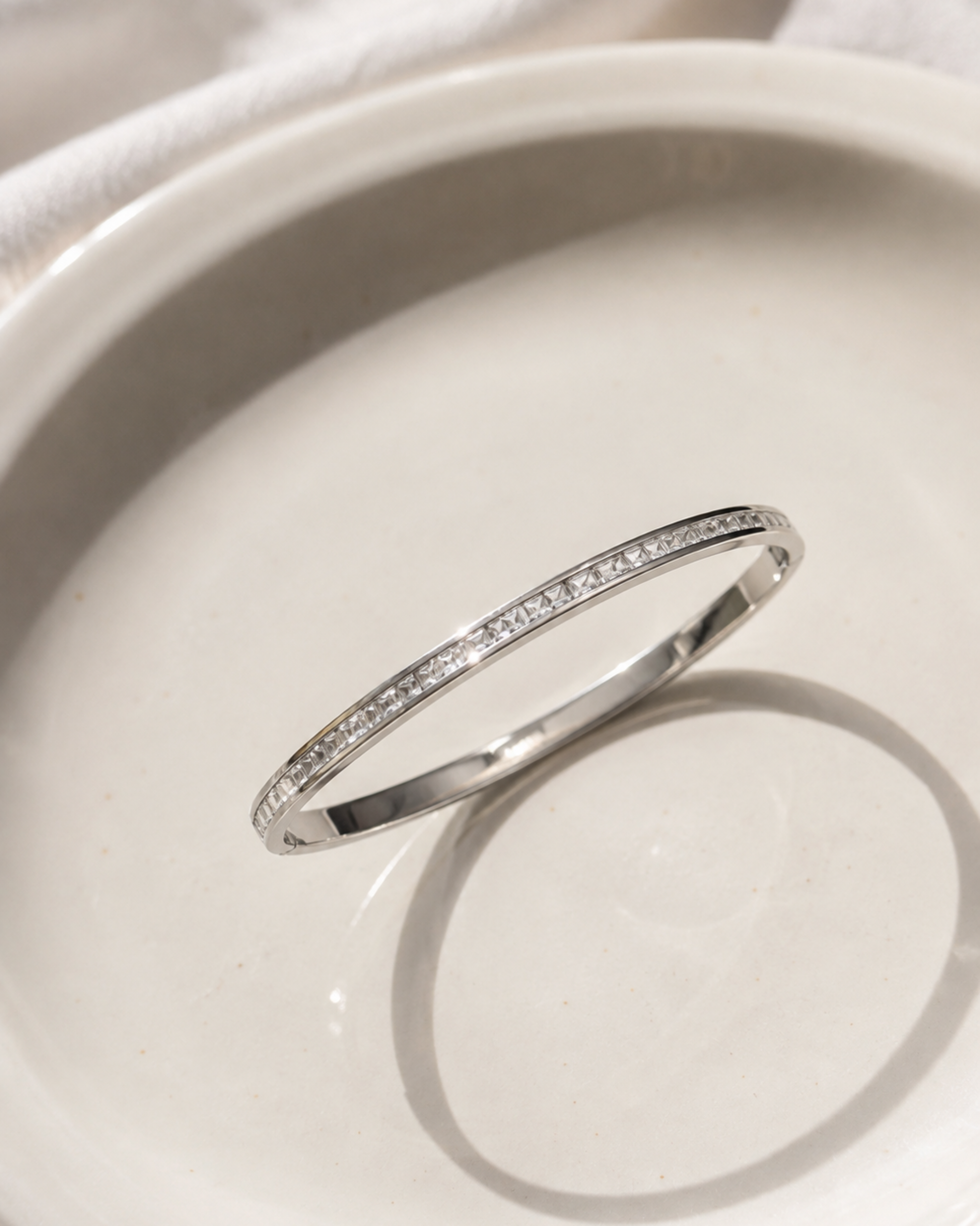 A thin silver band ring with a row of small clear stones, photographed resting on a white ceramic dish.