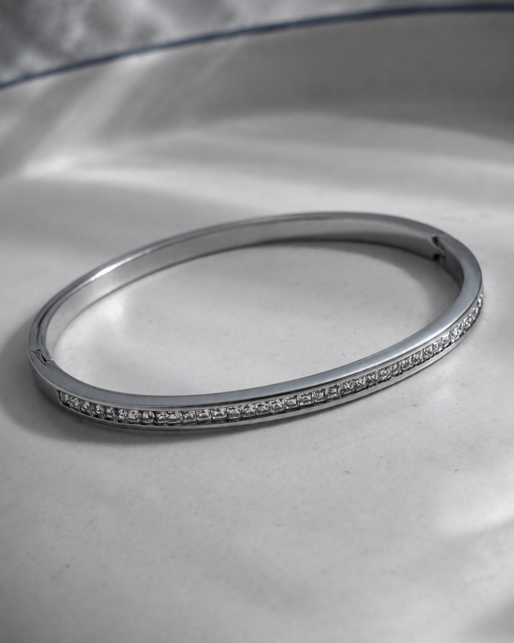 A slim silver bangle bracelet with a row of small clear stones along one side, photographed on a light gray satin-like surface.