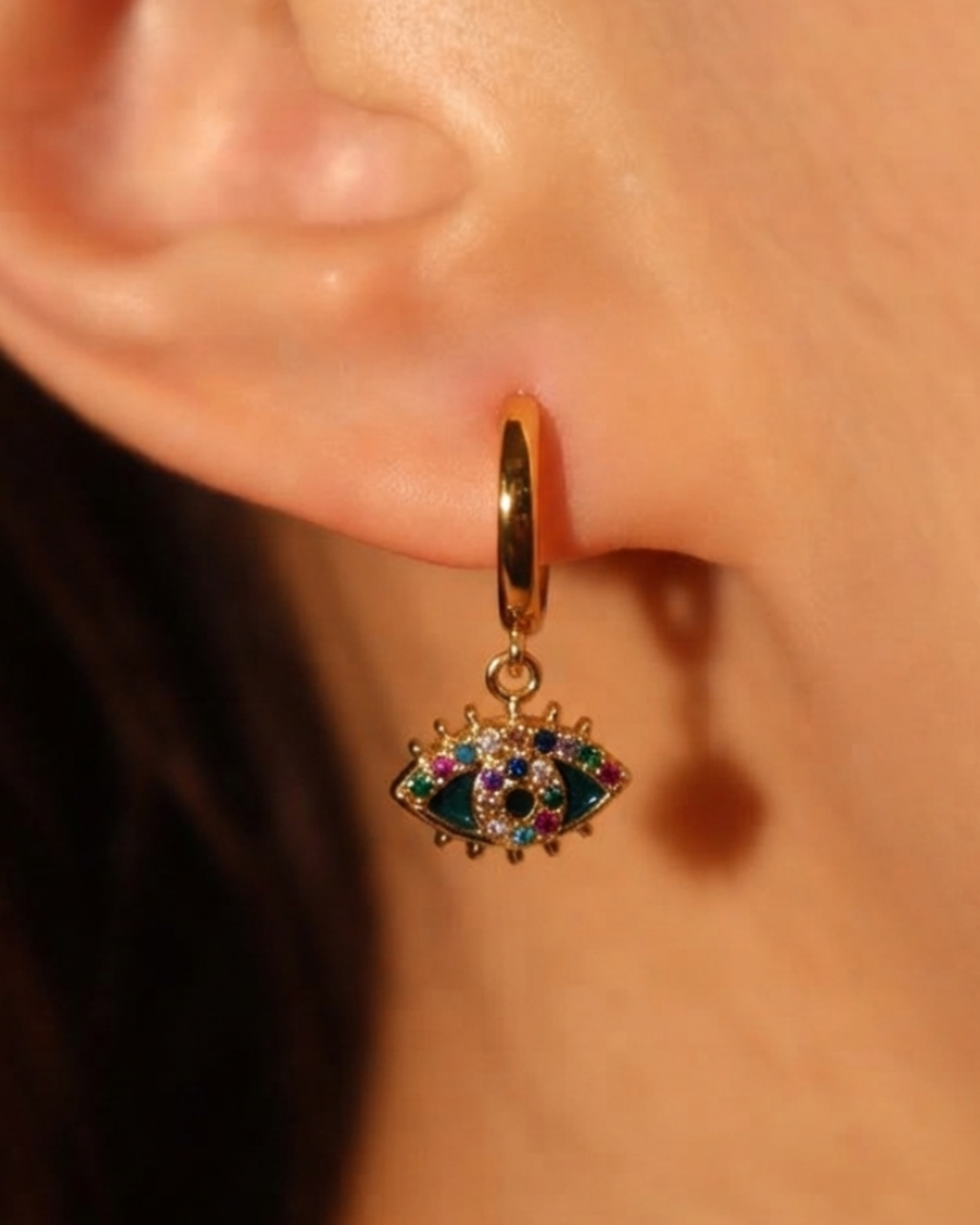 A gold huggie hoop earring with a dangling evil eye charm featuring green, blue, and pink stones, shown worn on a person's ear.