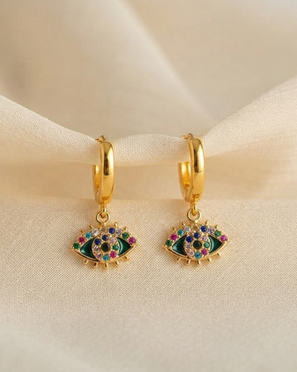 A pair of gold huggie hoop earrings with dangling evil eye charms, decorated with green enamel and colorful rhinestones, resting on a draped beige fabric background.
