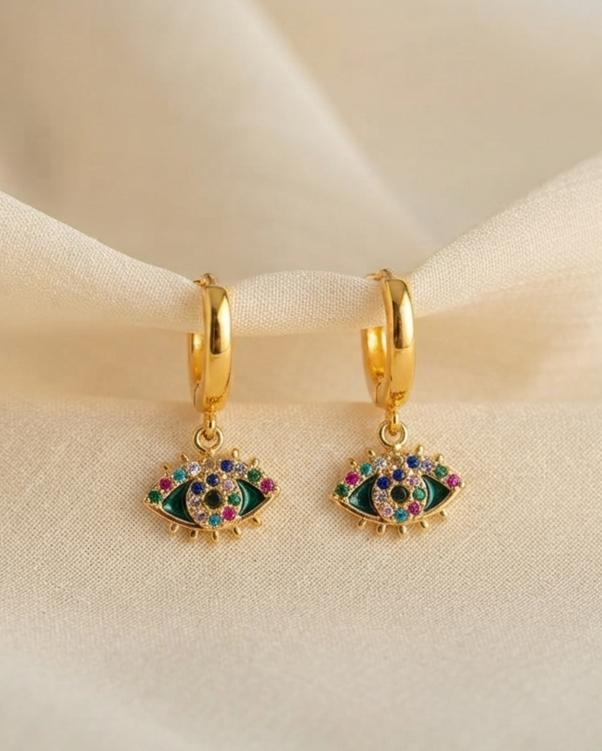 A pair of gold huggie hoop earrings with dangling evil eye charms, decorated with green enamel and colorful rhinestones, resting on a draped beige fabric background.