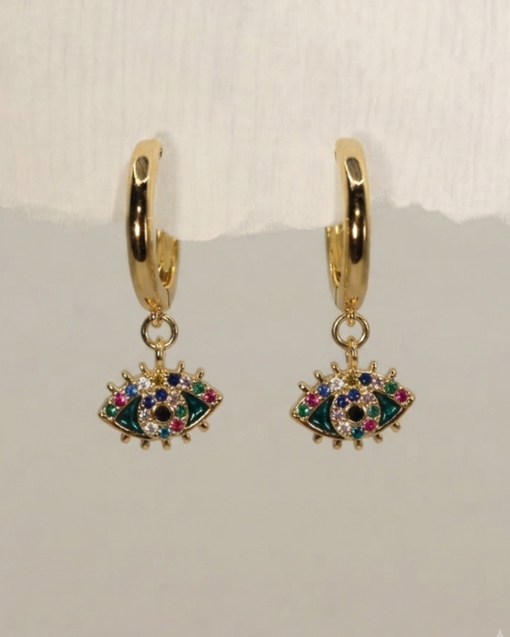 A pair of gold huggie hoop earrings with dangling evil eye charms embellished with green, blue, pink, and white rhinestones, displayed against a light gray background.