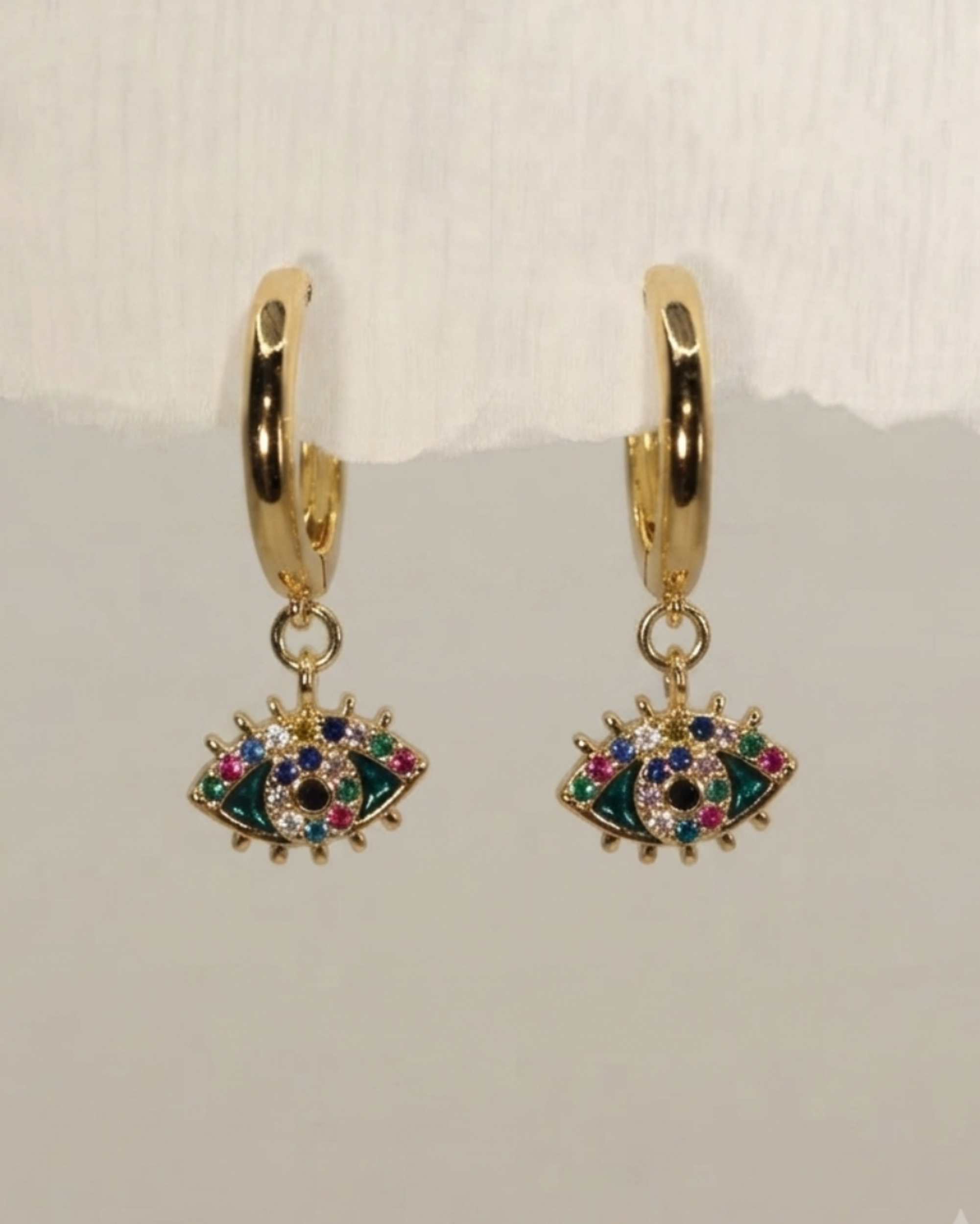 A pair of gold huggie hoop earrings with dangling evil eye charms embellished with green, blue, pink, and white rhinestones, displayed against a light gray background.