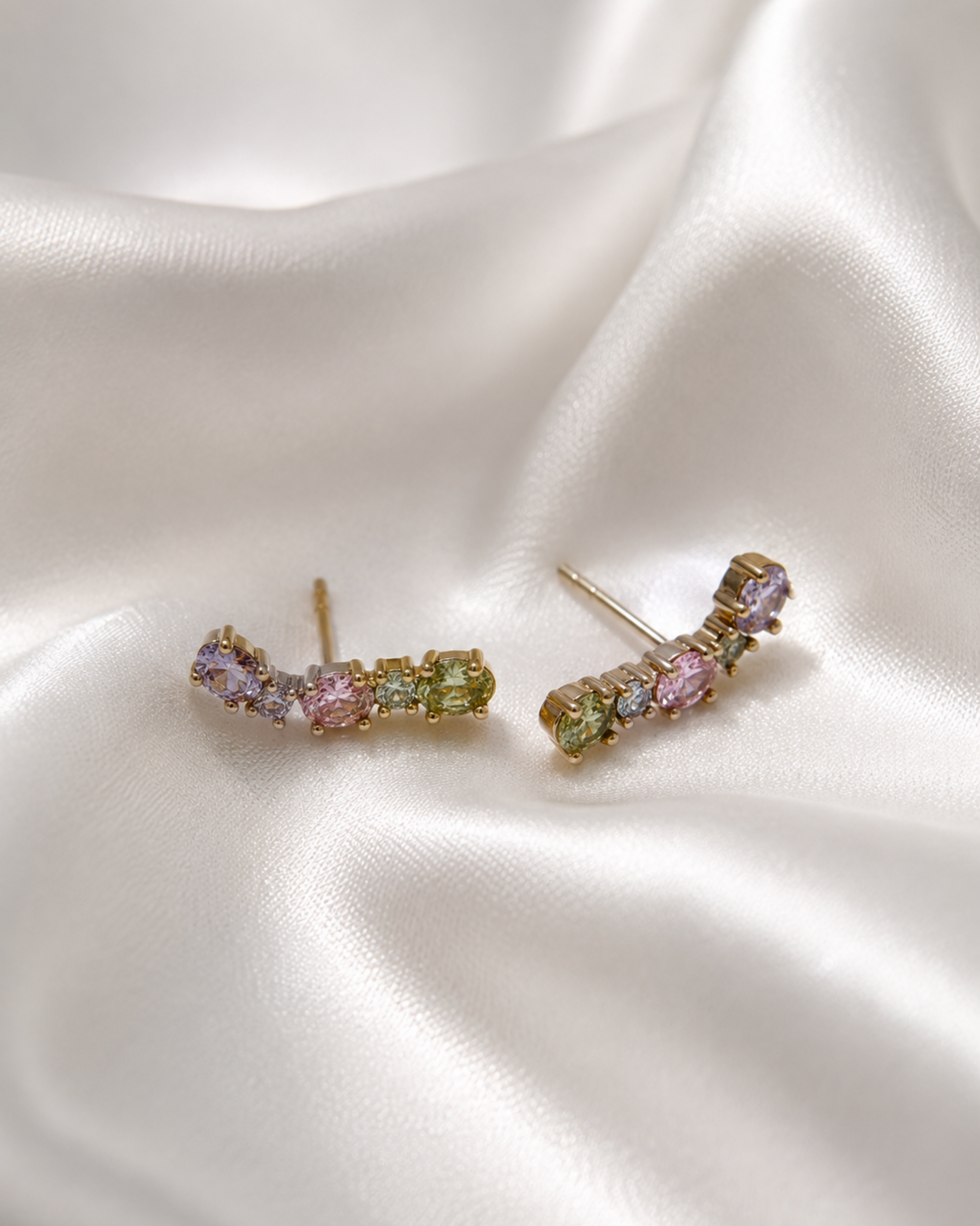 A pair of gold-toned curved stud earrings with small pastel gemstones in pink, green, and lavender, displayed on draped white satin fabric.