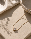 A delicate gold chain necklace with a small, clear heart-shaped gemstone pendant, styled on a textured beige linen surface with soft shadows.