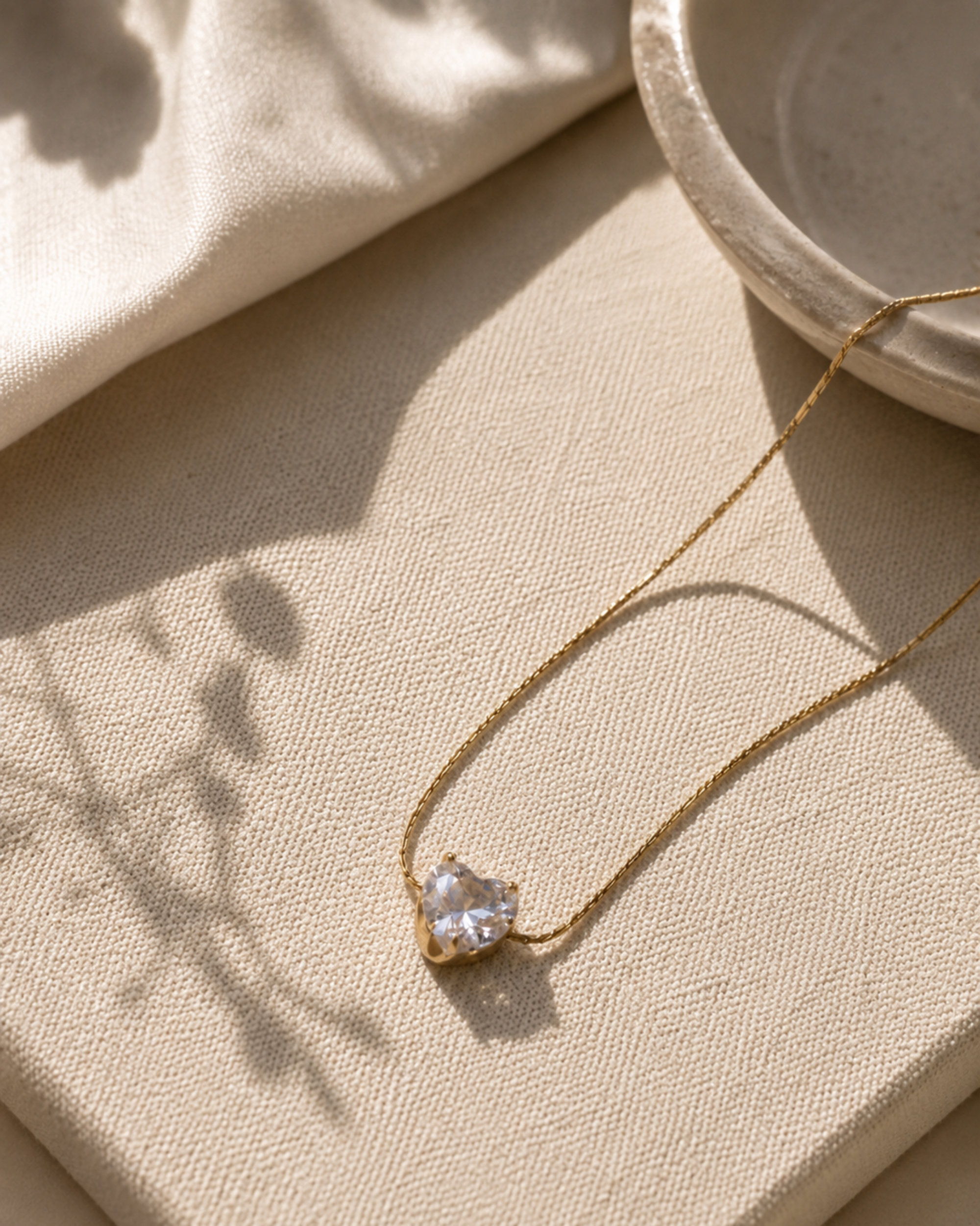 A delicate gold chain necklace with a small, clear heart-shaped gemstone pendant, styled on a textured beige linen surface with soft shadows.