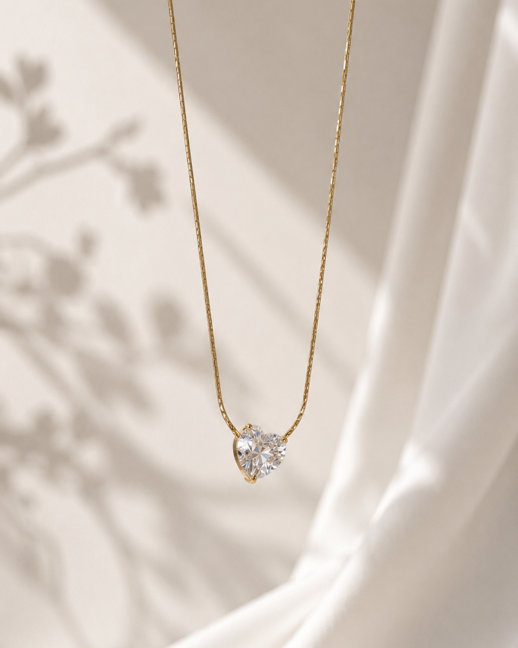 A delicate gold chain necklace with a small heart-shaped clear gemstone pendant, displayed against a soft cream-colored draped fabric background with soft shadows.