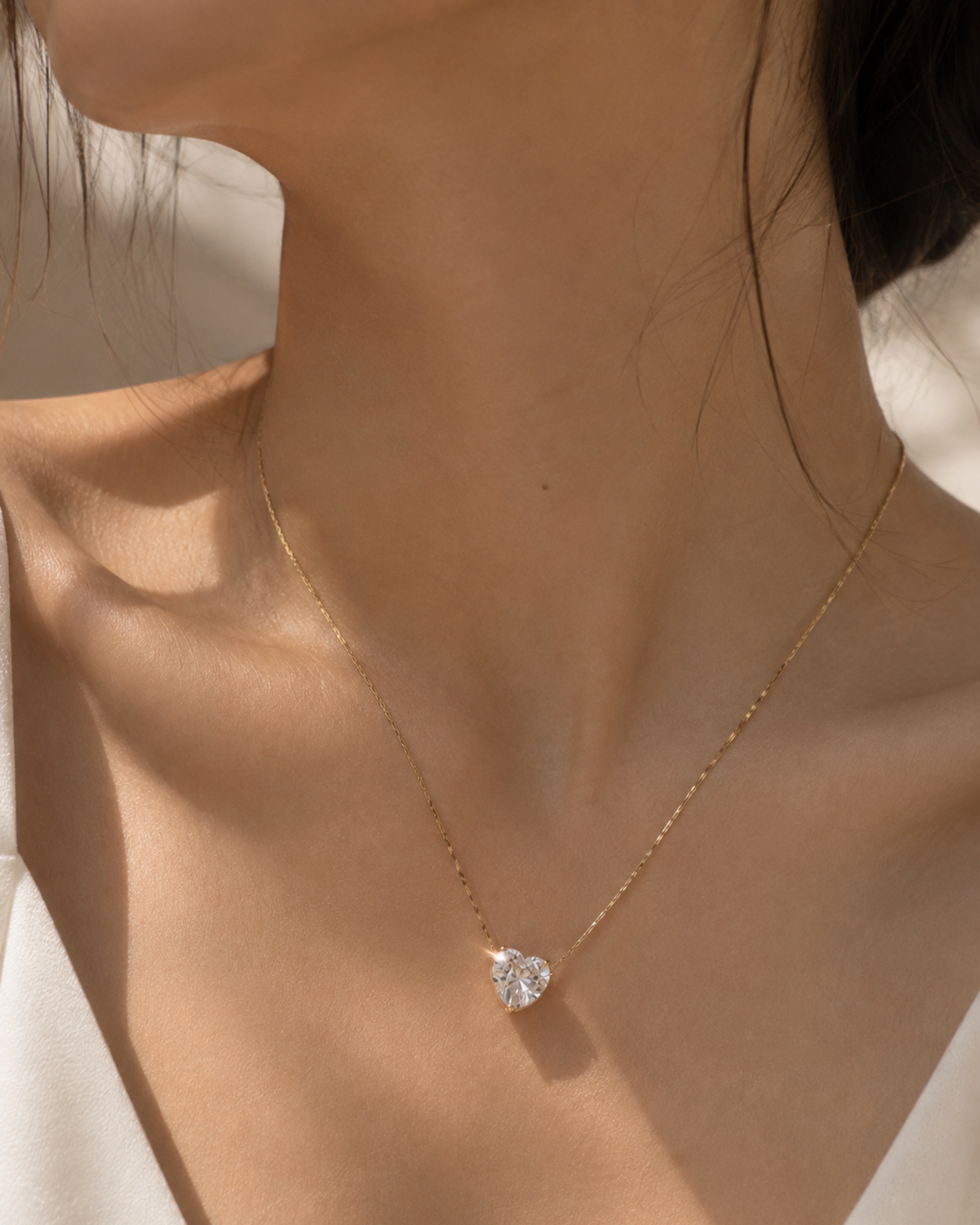 A close-up of a model's neck and chest wearing a delicate gold chain necklace with a clear heart-shaped gemstone pendant.