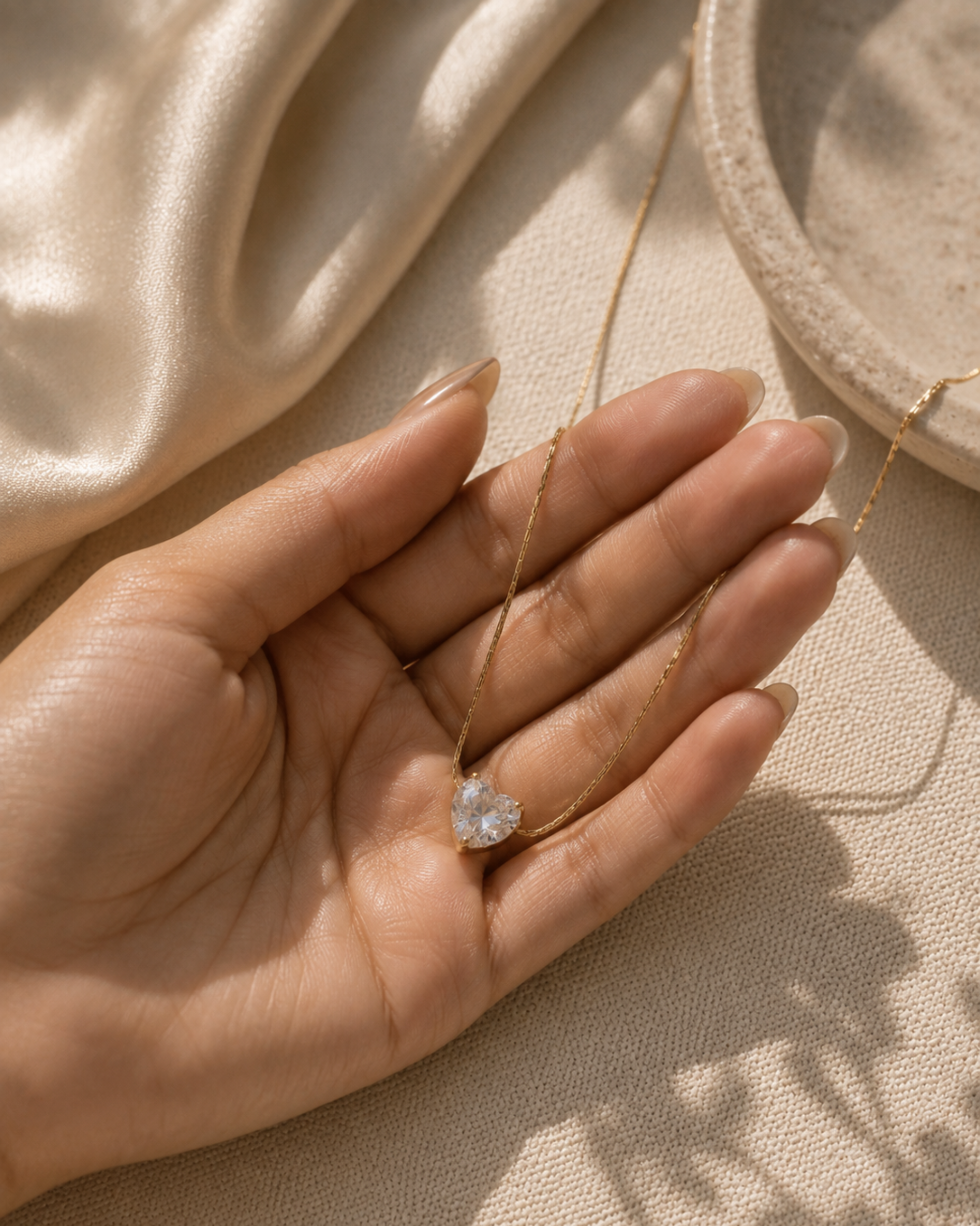 A delicate gold necklace with a heart-shaped clear gemstone pendant, held in the palm of a hand against a warm beige fabric background with a cream satin cloth and a textured dish.
