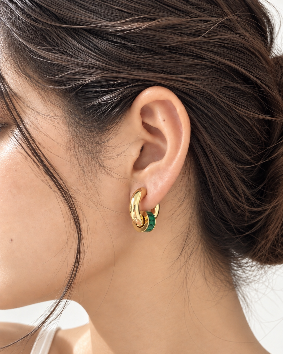 A close-up side profile of a woman wearing a pair of gold huggie hoop earrings with a thin green accent stripe, set against a clean white background.