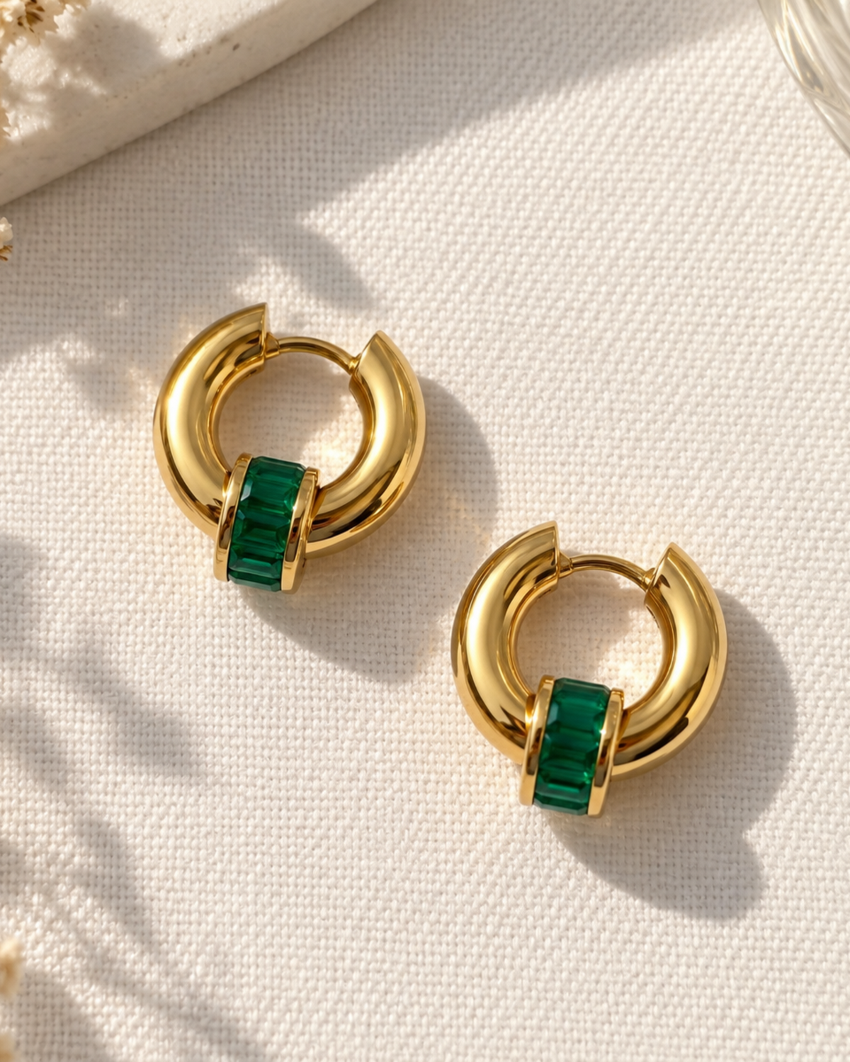A pair of gold crescent hoop earrings with a rectangular green crystal accent, displayed on a textured cream fabric with dried flowers.