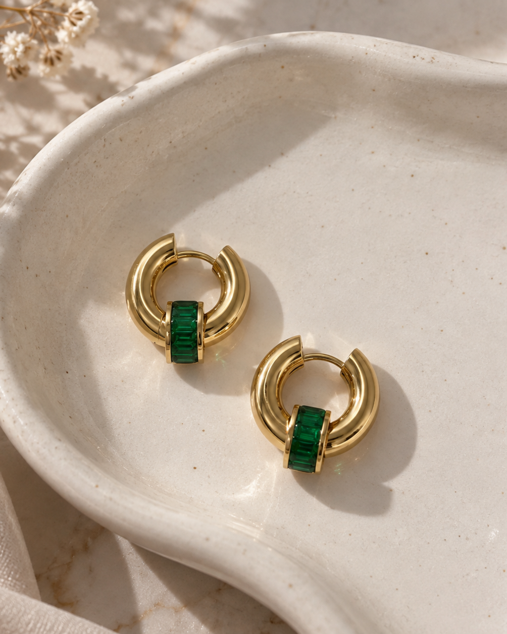 A pair of gold huggie hoop earrings with a central green textured bar, displayed on a white ceramic dish with dried flowers.