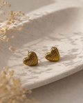 A pair of textured gold heart stud earrings resting on a light beige ceramic dish with dried flowers in the foreground.