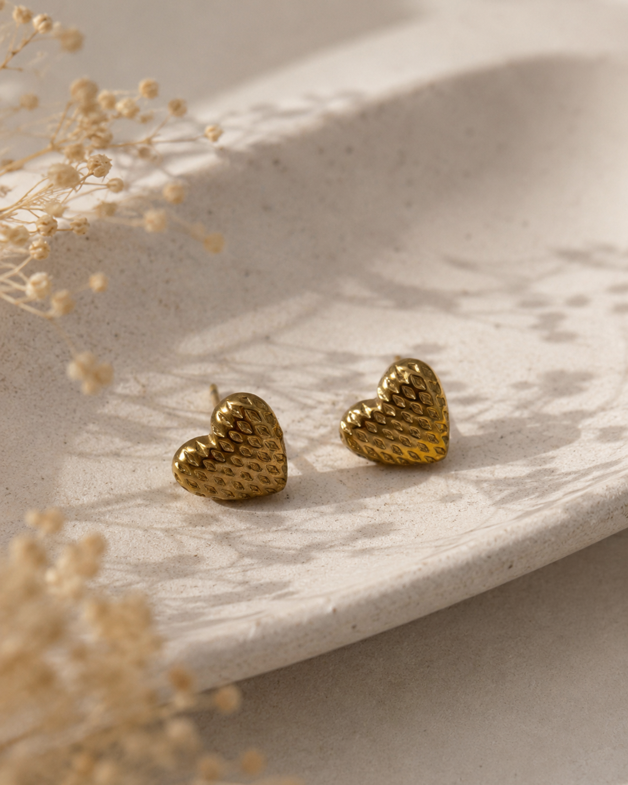 A pair of textured gold heart stud earrings resting on a light beige ceramic dish with dried flowers in the foreground.