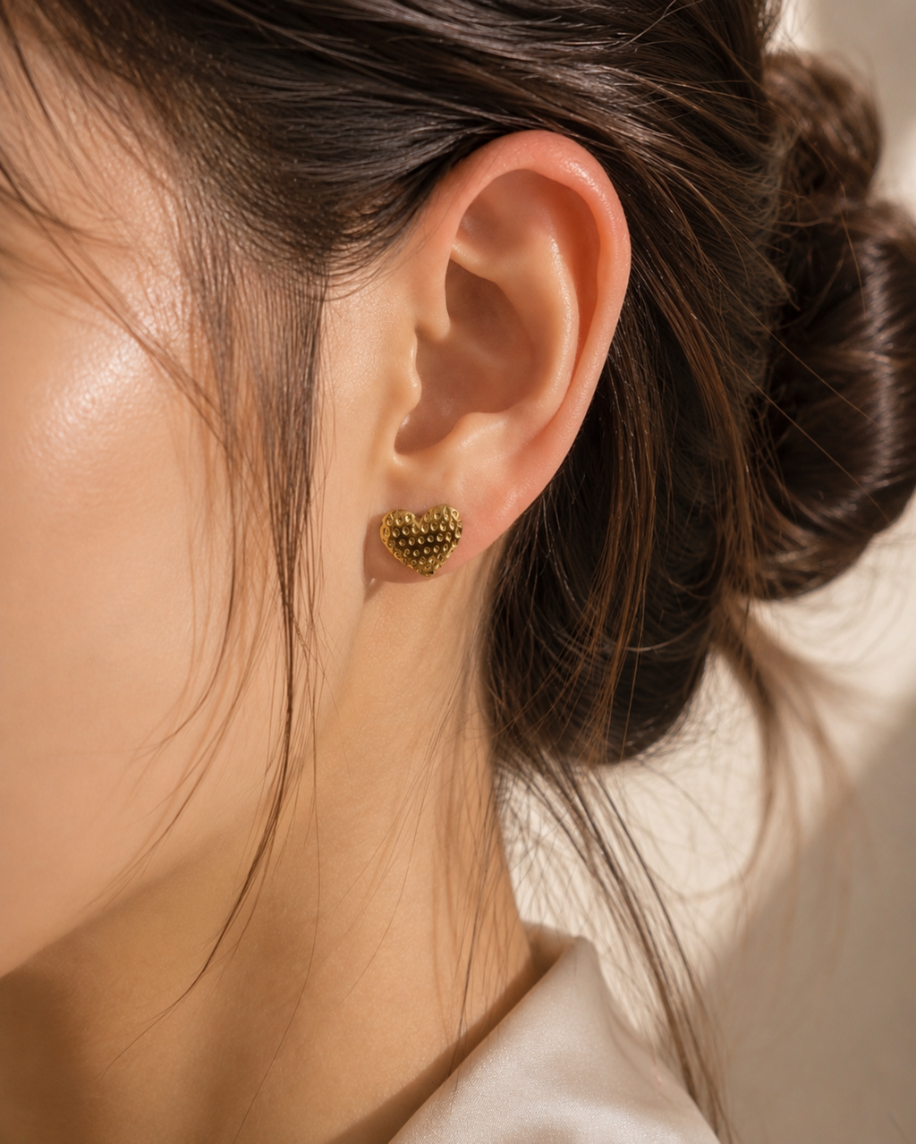 A close-up of a model's ear wearing a small textured gold heart-shaped stud earring.