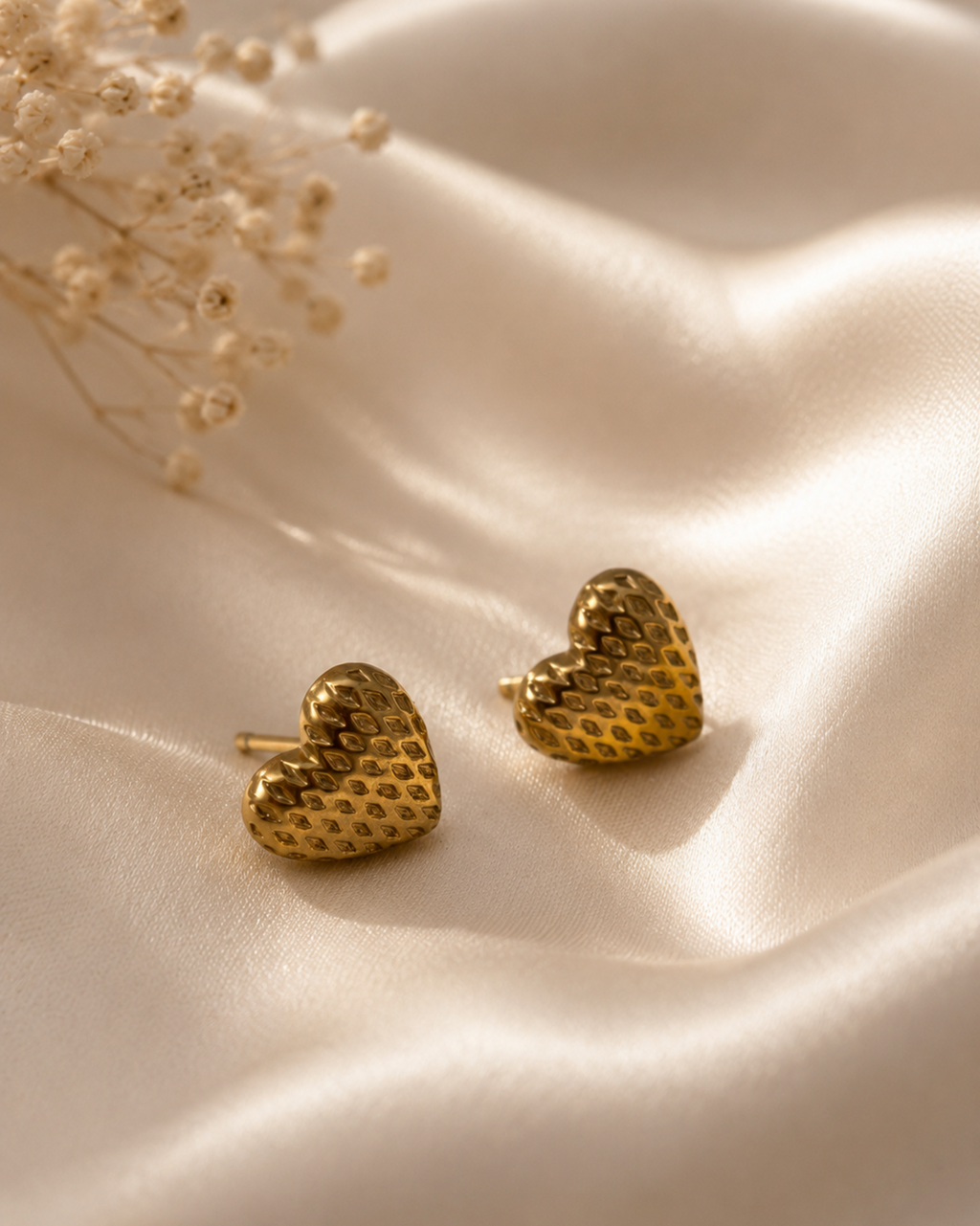 Two small gold heart-shaped stud earrings with a textured, dotted surface finish, displayed on draped cream satin fabric with dried baby's breath flowers in the corner.