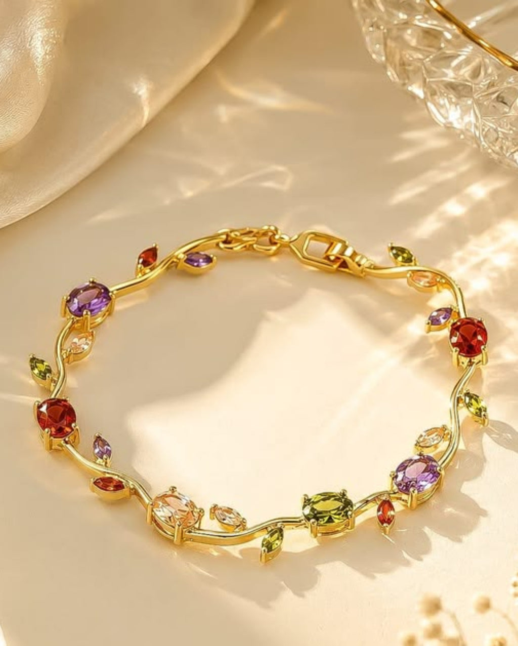 A gold vine-style bracelet adorned with colorful gemstones, including purple, red, green, and peach, displayed on a warm cream satin surface with soft shadows.