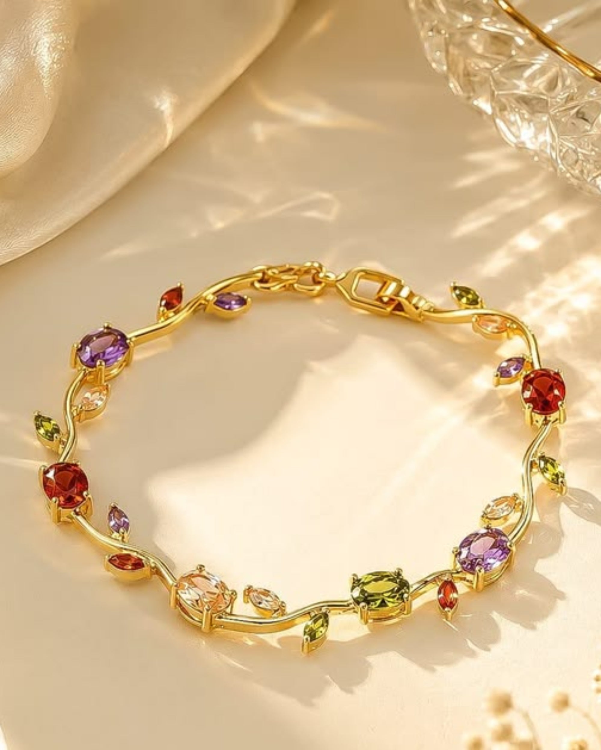 A gold vine-style bracelet adorned with colorful gemstones, including purple, red, green, and peach, displayed on a warm cream satin surface with soft shadows.