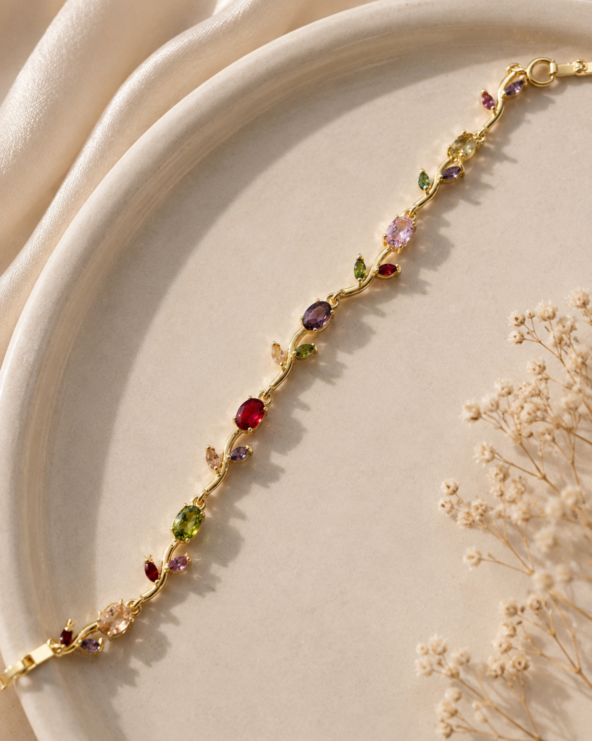A delicate gold vine bracelet with colorful gemstone leaves, displayed on a cream-colored ceramic plate with dried flowers and soft cream fabric in the background.