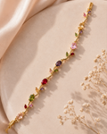 A gold floral bracelet adorned with colorful gemstones, displayed diagonally on a beige satin surface with dried baby's breath flowers.