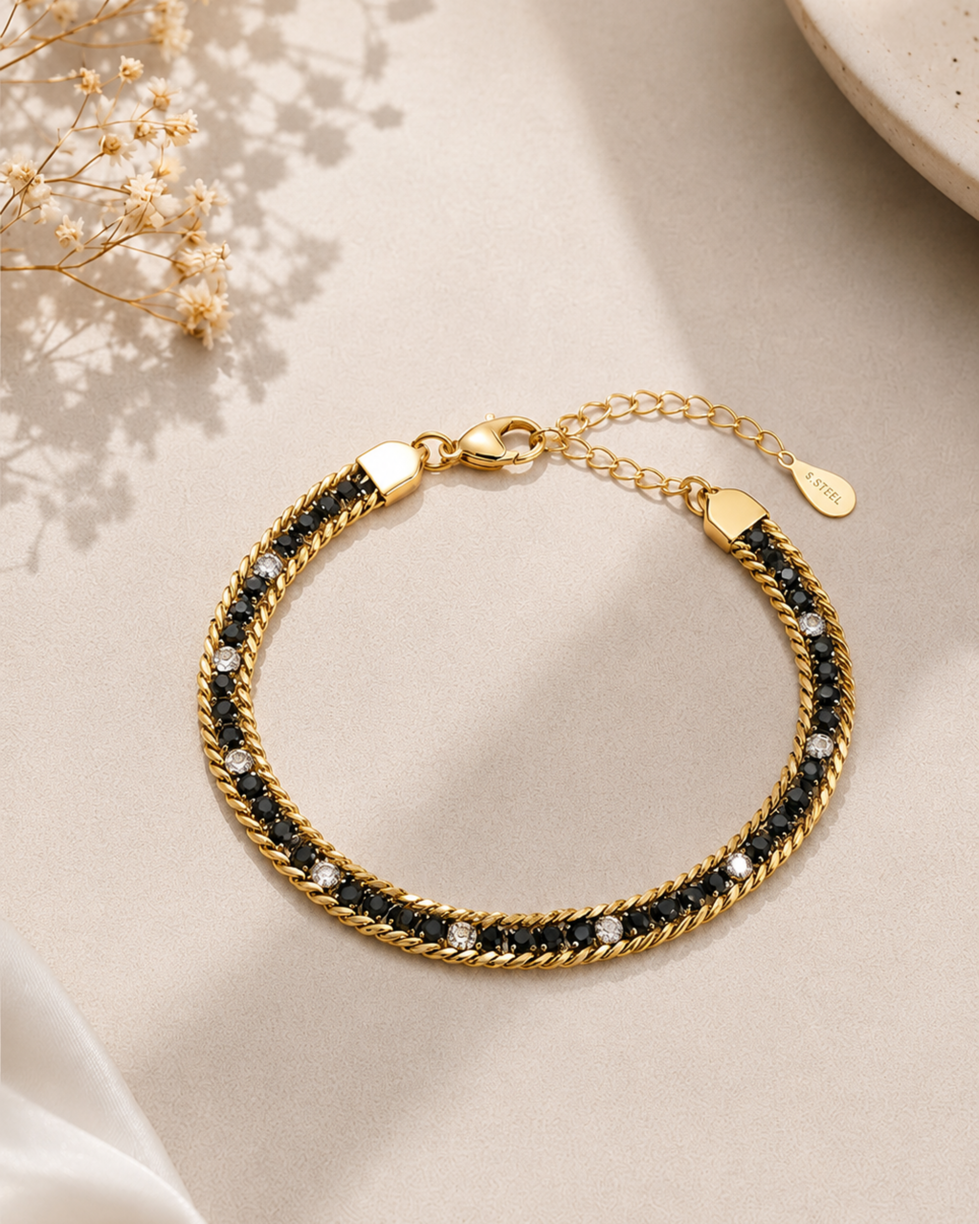 A gold-toned rope chain bracelet with black woven inlay and small clear stones, displayed on a light beige surface with soft shadows and dried flowers in the corner.