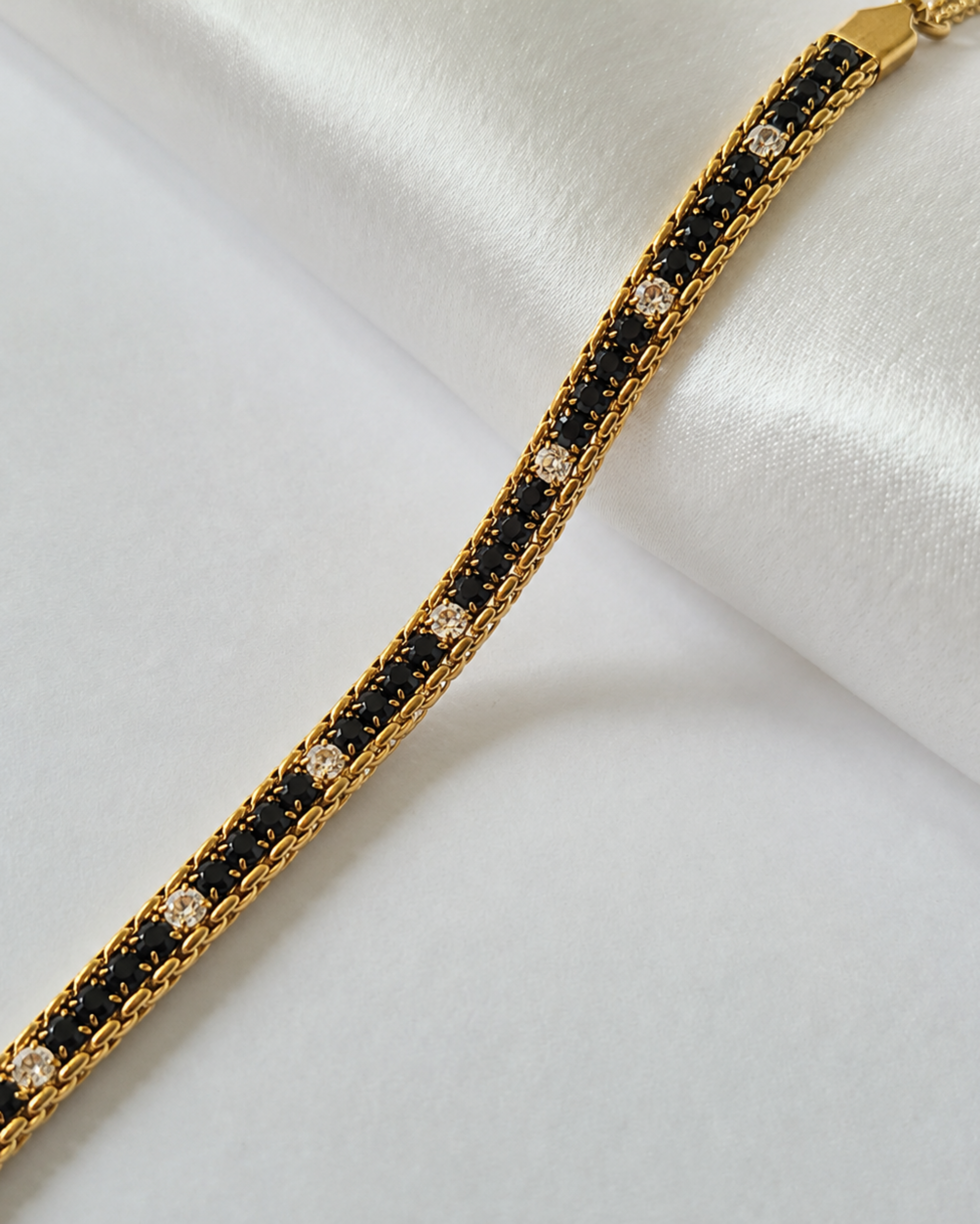 A gold-toned tennis bracelet featuring alternating rows of small black stones and clear cubic zirconia stones, displayed on a white satin fabric background.