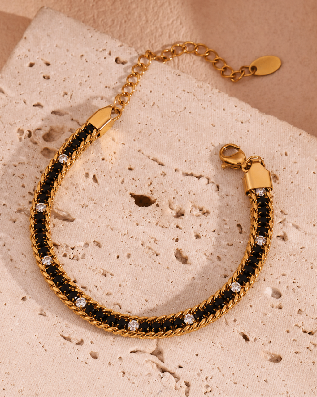 A gold chain bracelet with a black woven center and small clear rhinestones, displayed on a textured beige stone surface.