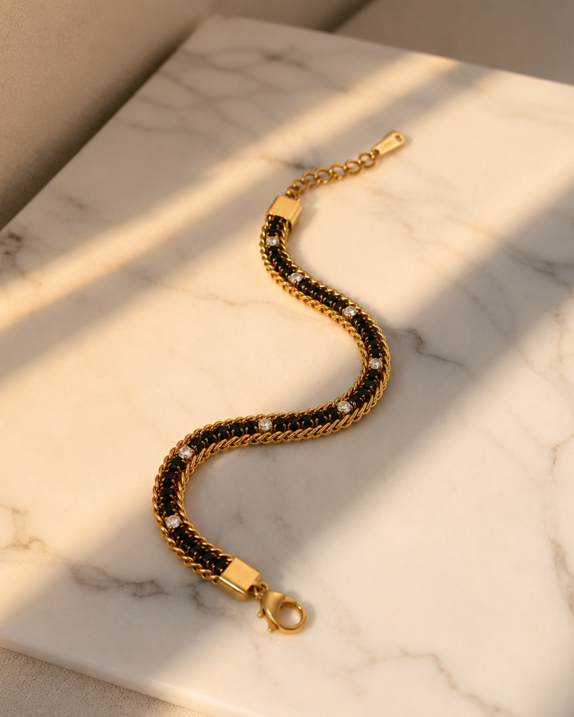 A gold chain bracelet with black woven inlay and small clear stones, displayed on a white marble surface with warm sunlight and shadows.