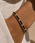 A close-up of a gold bangle bracelet with black stones and clear crystal accents, worn on a person's wrist.