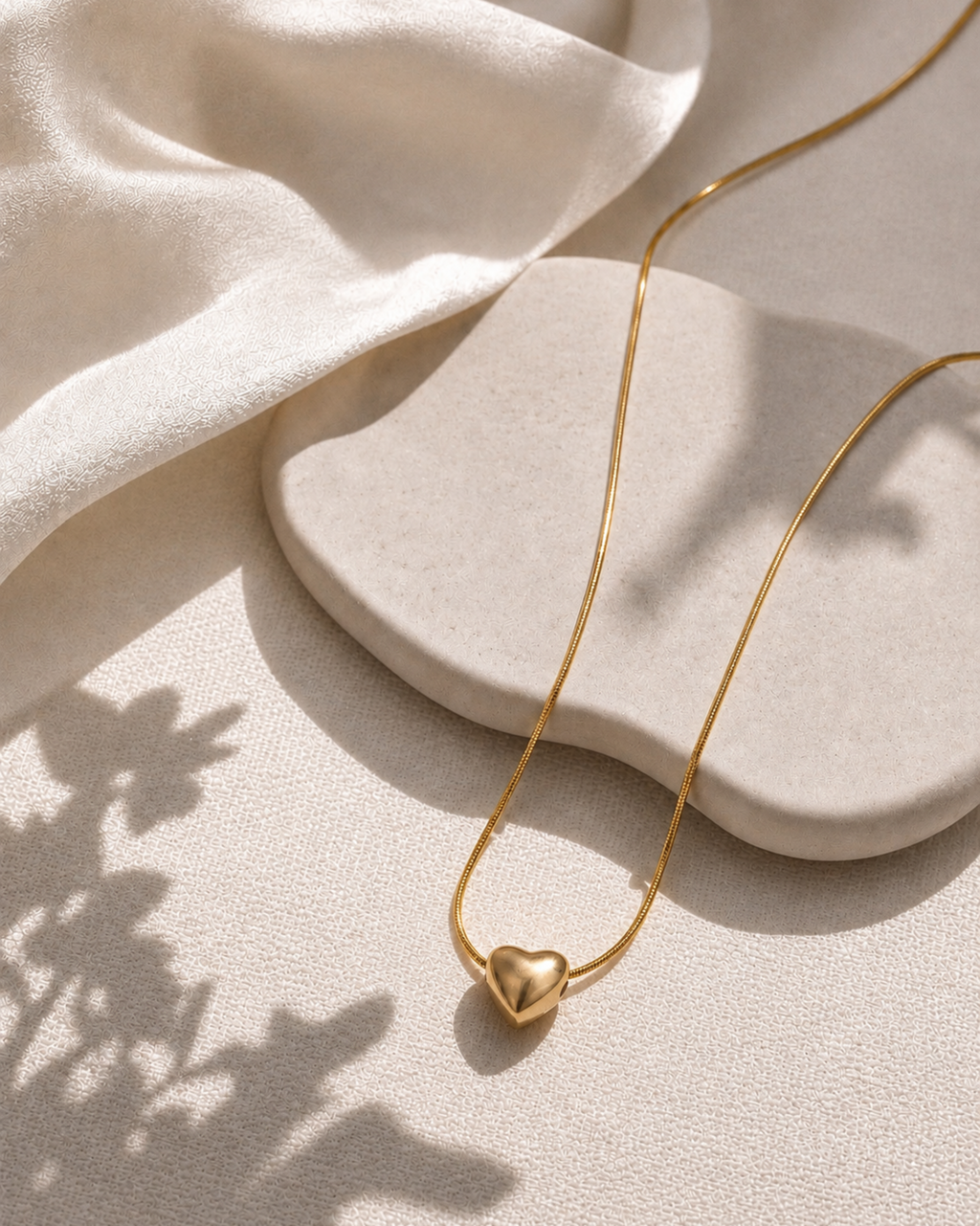 A delicate gold chain necklace with a small polished gold heart pendant, styled on a textured beige surface with draped cream fabric and soft shadows.