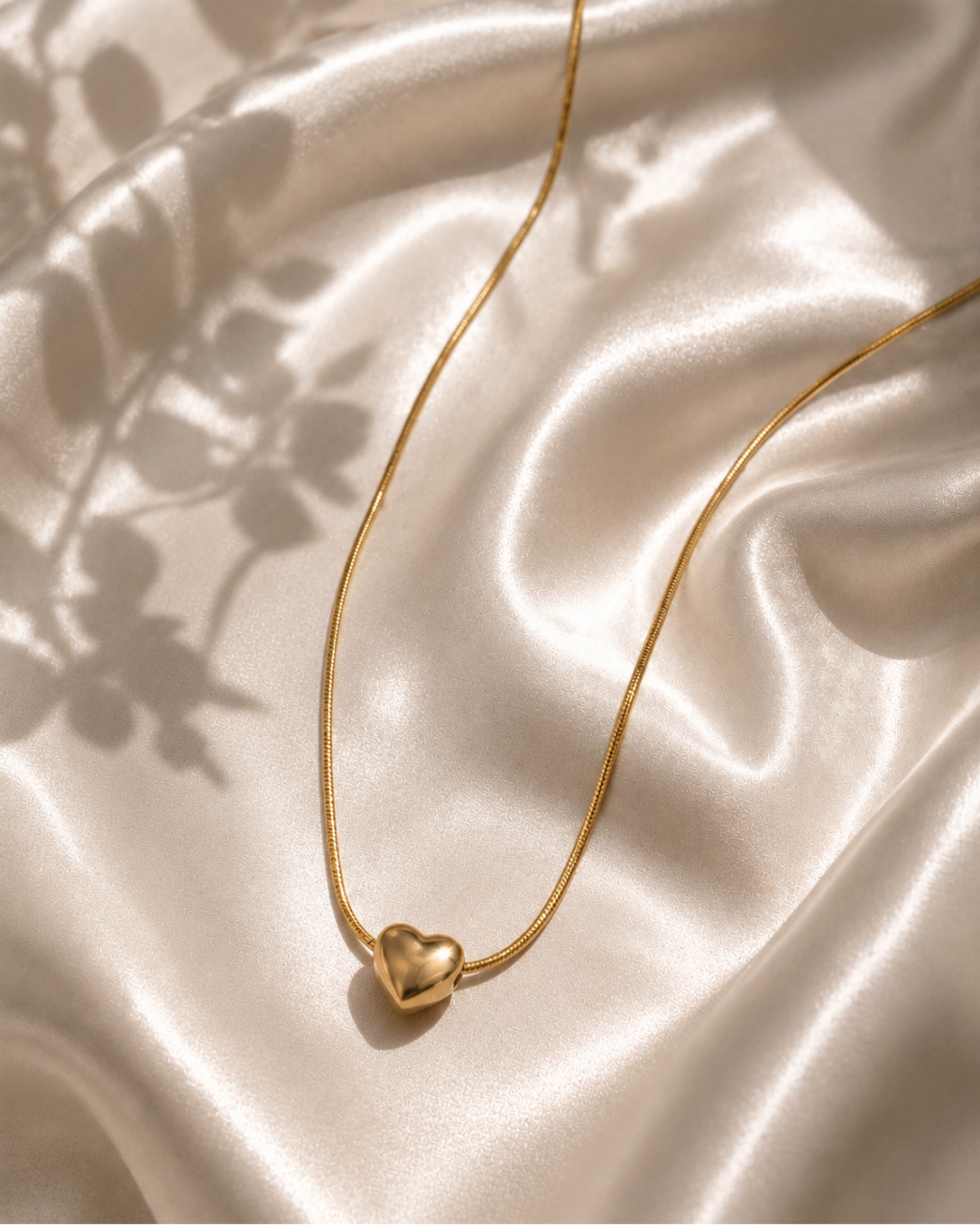 A delicate gold chain necklace with a small polished heart pendant, draped elegantly on draped champagne satin fabric with soft leaf shadows.