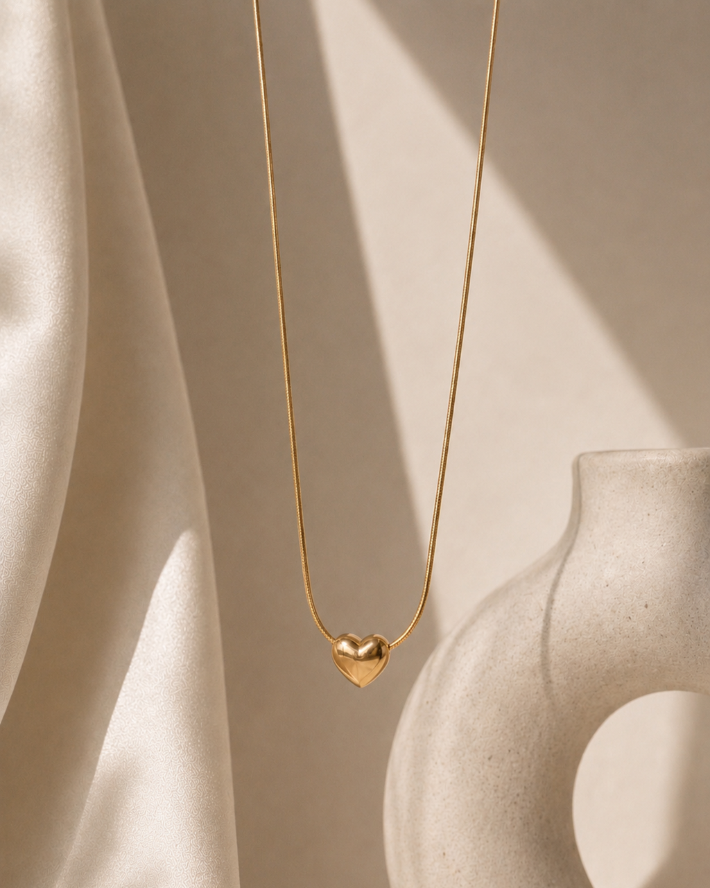 A delicate gold chain necklace with a small, polished gold heart pendant, styled against a warm beige background with draped satin fabric and a ceramic vase.