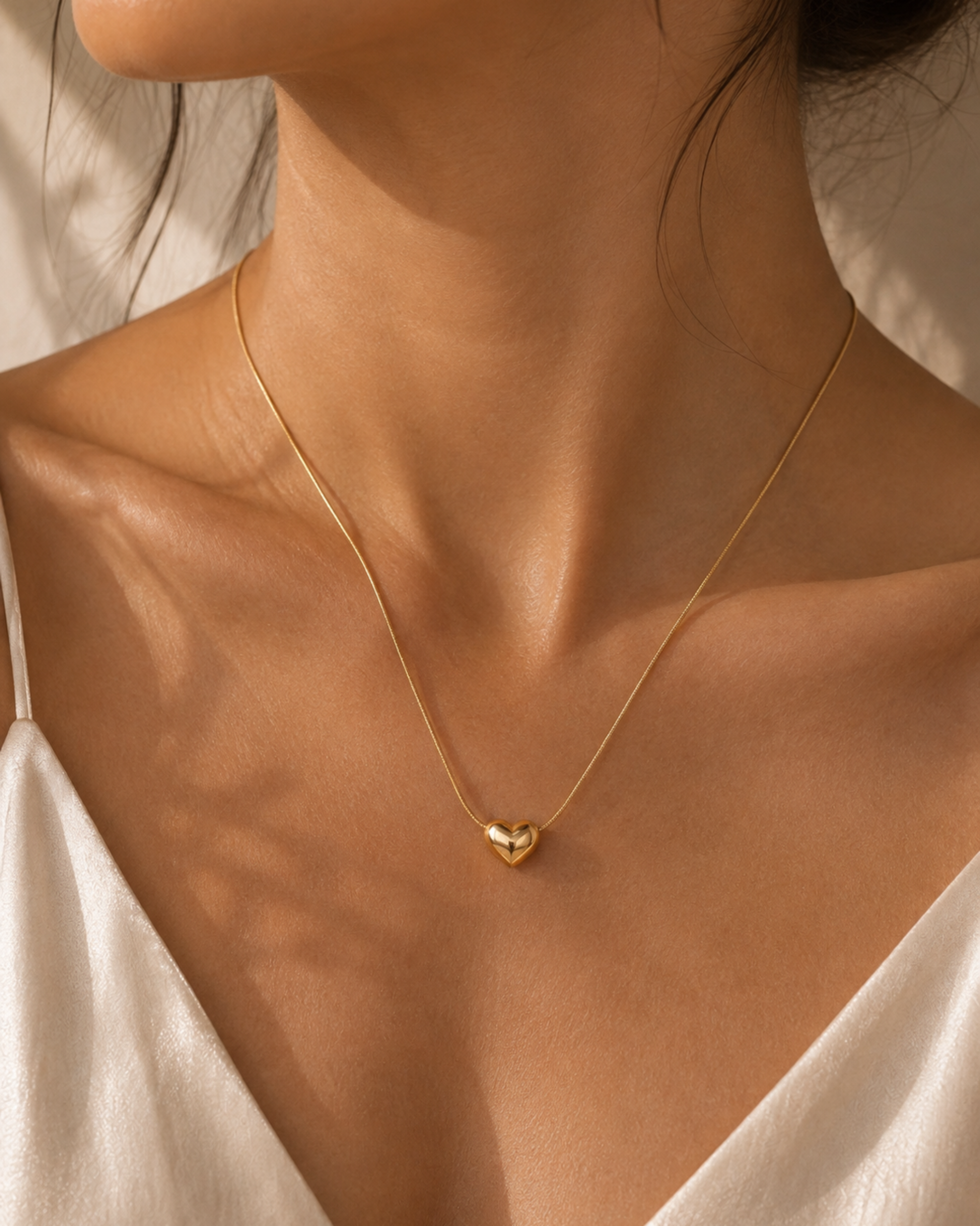 A close-up of a model's neck and upper chest wearing a delicate gold chain necklace with a small, polished gold heart pendant, styled with a white satin top.