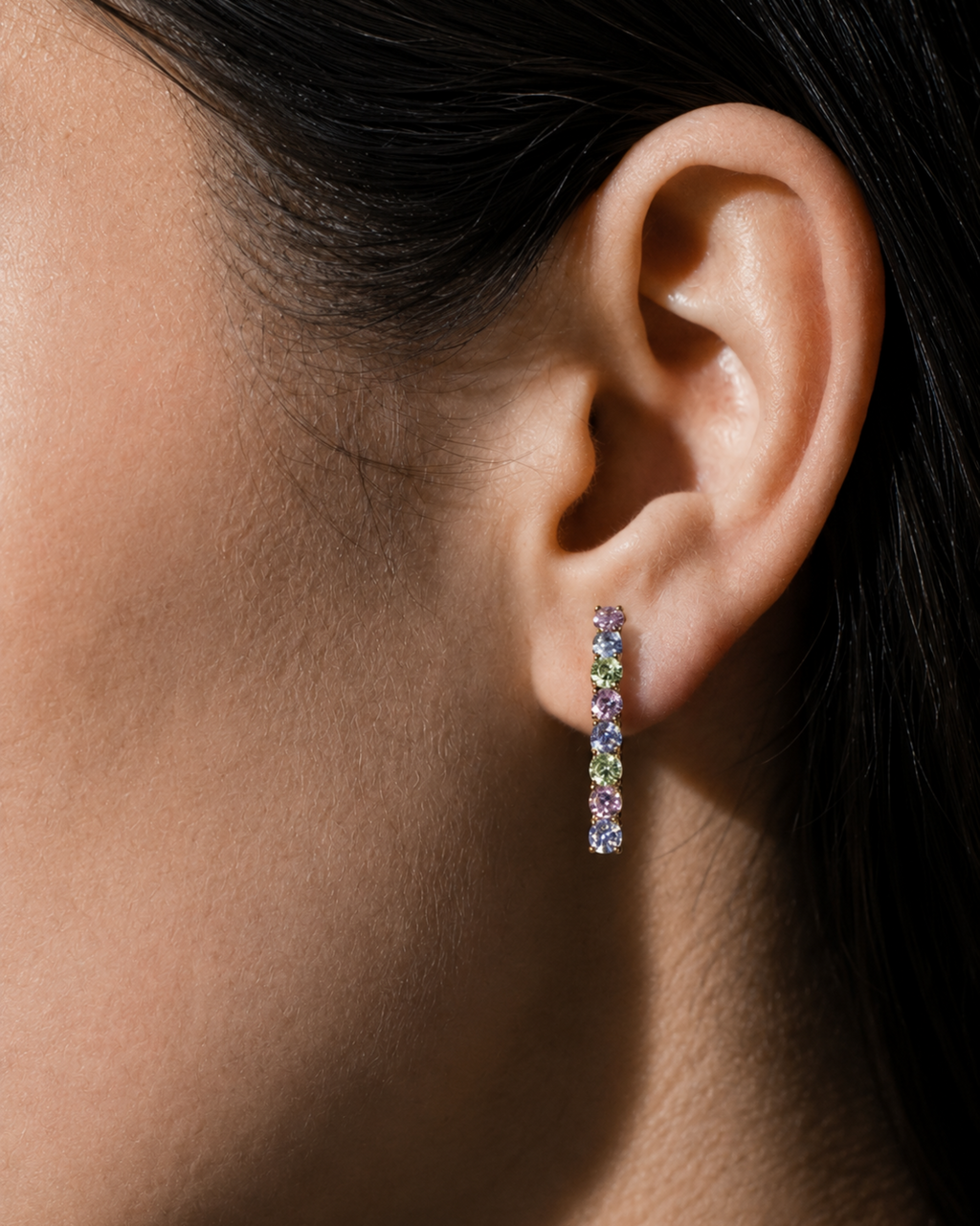 A close-up of a model's ear wearing a slim vertical dangle earring set with small multicolored gemstones.