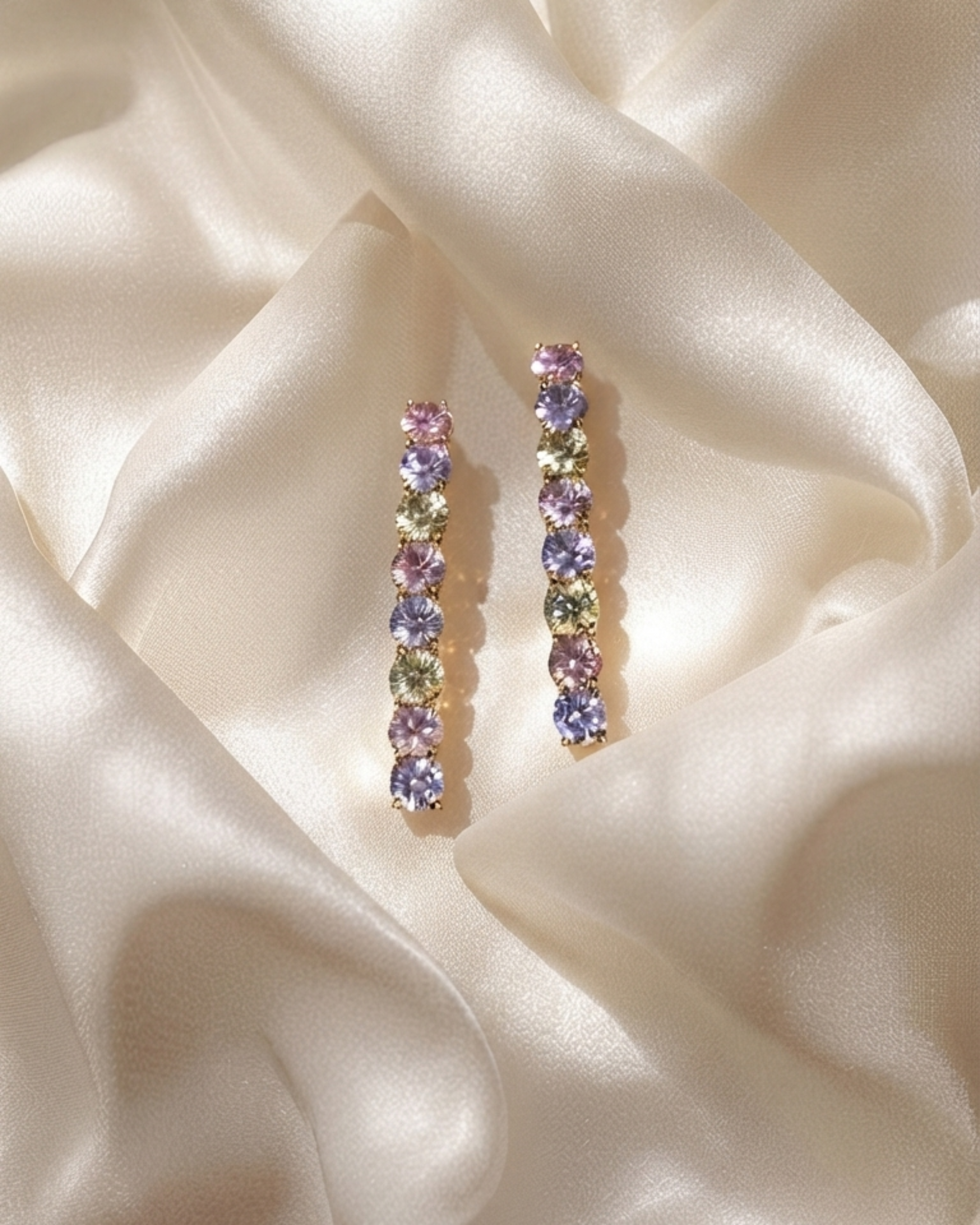 A pair of gold-toned drop earrings with a row of round, multicolored gemstones, displayed on draped cream satin fabric.