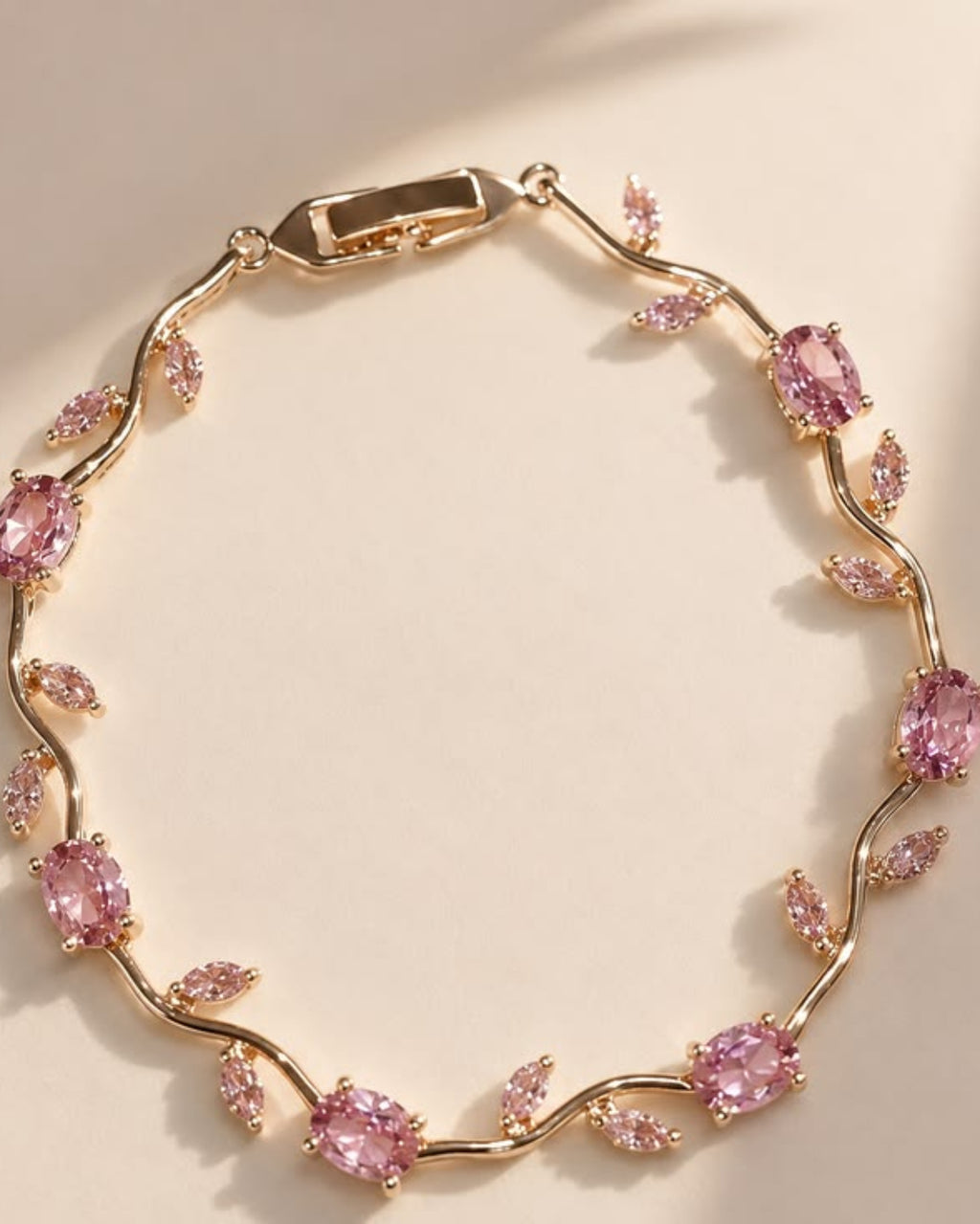 A gold-plated vine bracelet adorned with pink gemstones, displayed on a warm beige background with soft diagonal shadows.