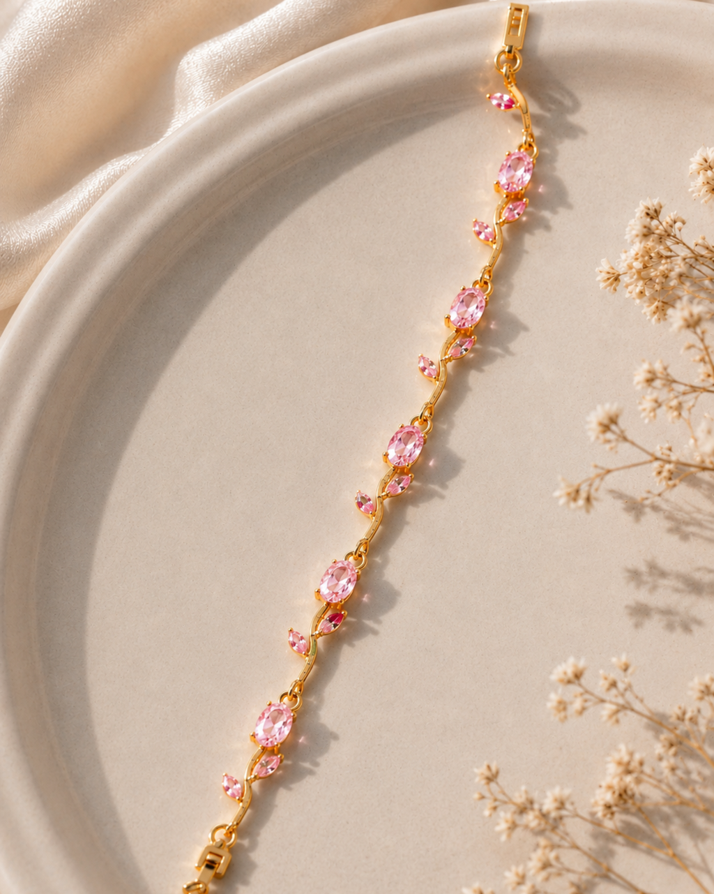 A gold-toned bracelet with multiple oval pink gemstones, styled diagonally on a beige ceramic plate with soft cream fabric and dried flowers.