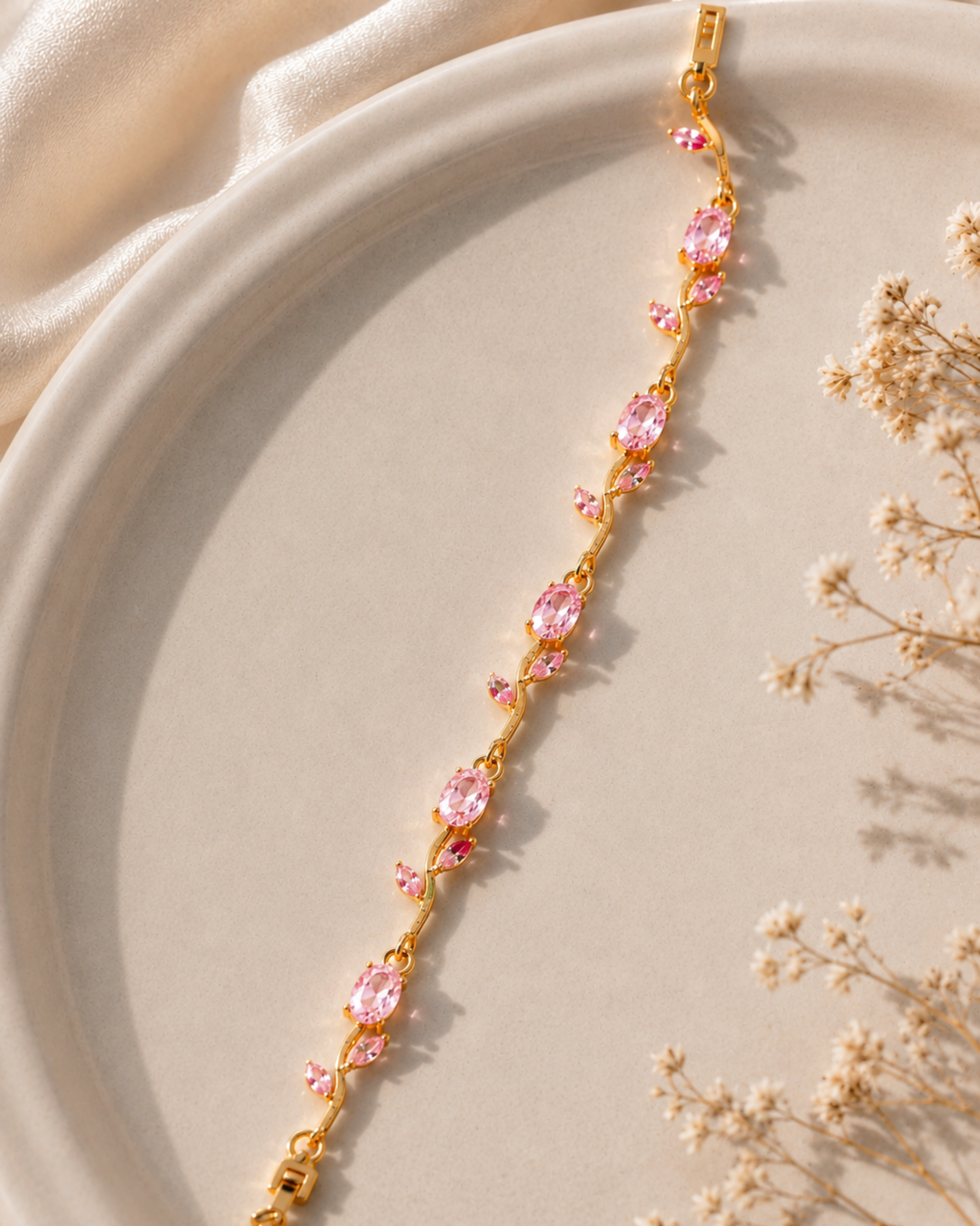 A gold-toned bracelet with multiple oval pink gemstones, styled diagonally on a beige ceramic plate with soft cream fabric and dried flowers.