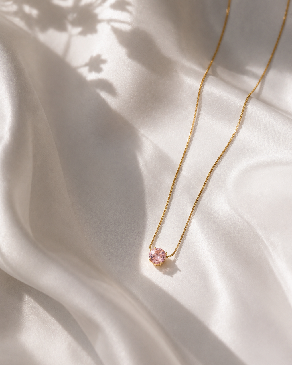 A delicate gold chain necklace with a small pink flower-shaped pendant, laid out on draped white satin fabric with soft shadows.