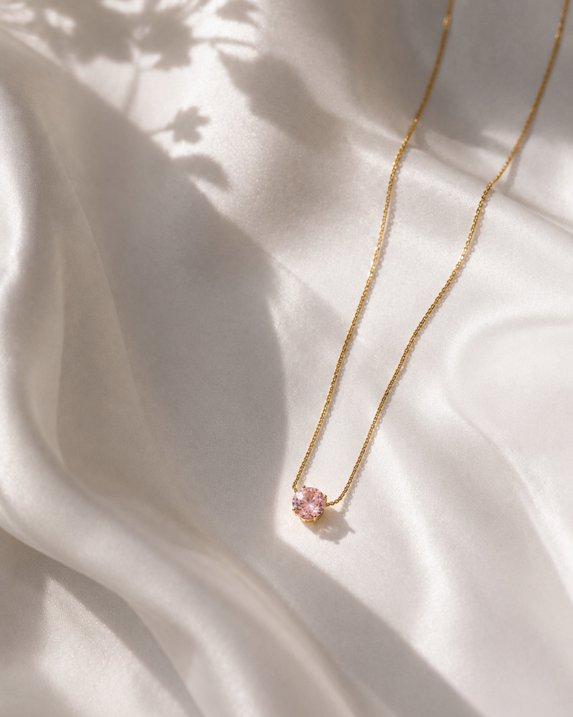A delicate gold chain necklace with a small pink flower-shaped pendant, laid out on draped white satin fabric with soft shadows.