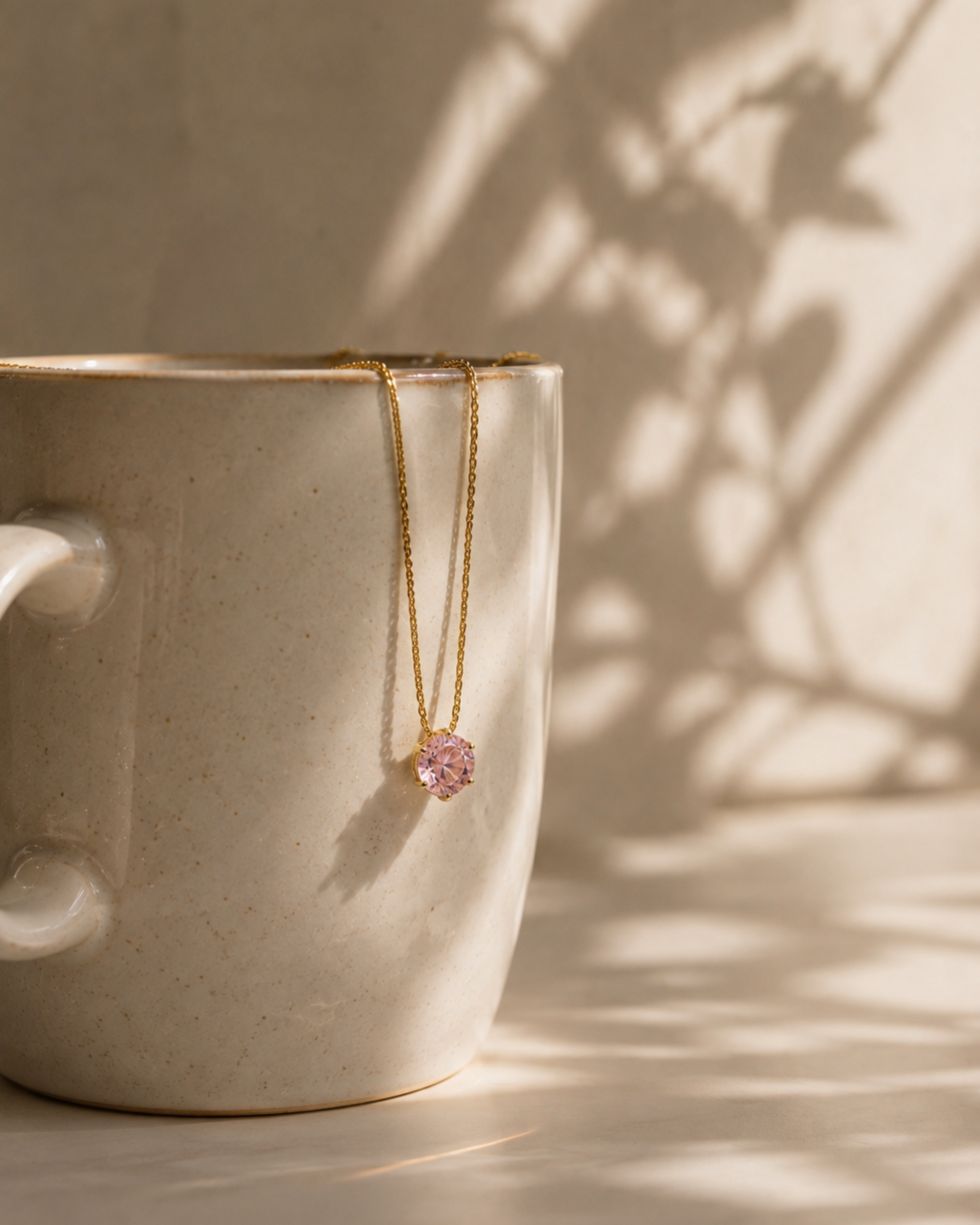A delicate gold necklace with a small pink gemstone pendant draped over the rim of a speckled beige ceramic mug, set against a warm beige wall with soft leaf shadows.