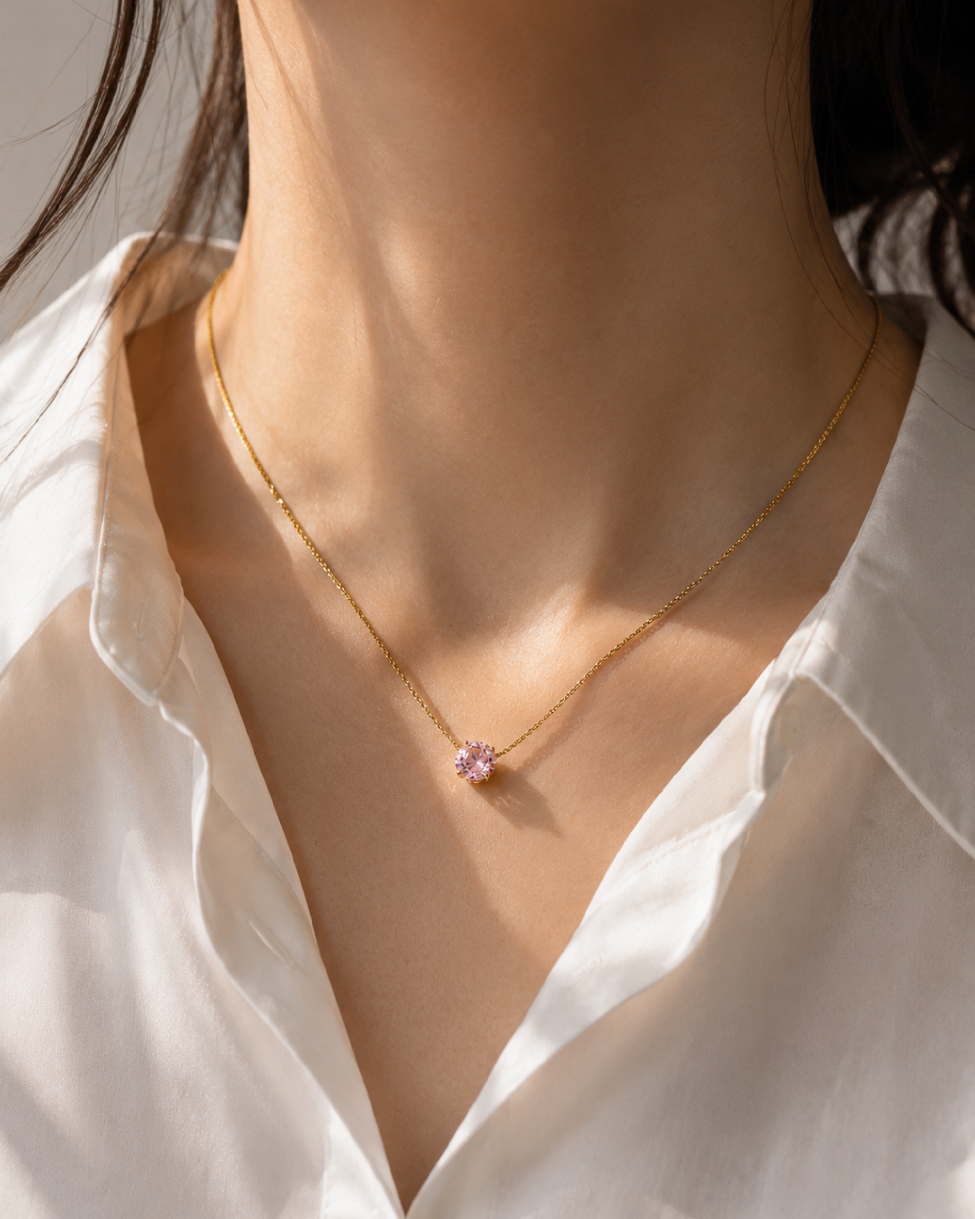 A close-up of a model wearing a delicate gold chain necklace with a small, round, light pink gemstone pendant, styled with a white collared shirt.