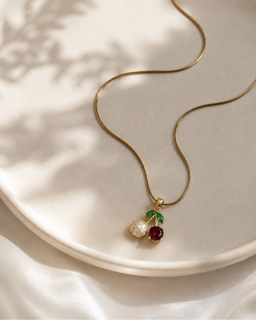 A gold snake chain necklace with a cherry pendant, resting on a light beige ceramic plate with soft leaf shadows. The pendant features a gold-toned metal setting with a clear pavé stone, a deep red gemstone, and a small green stone.